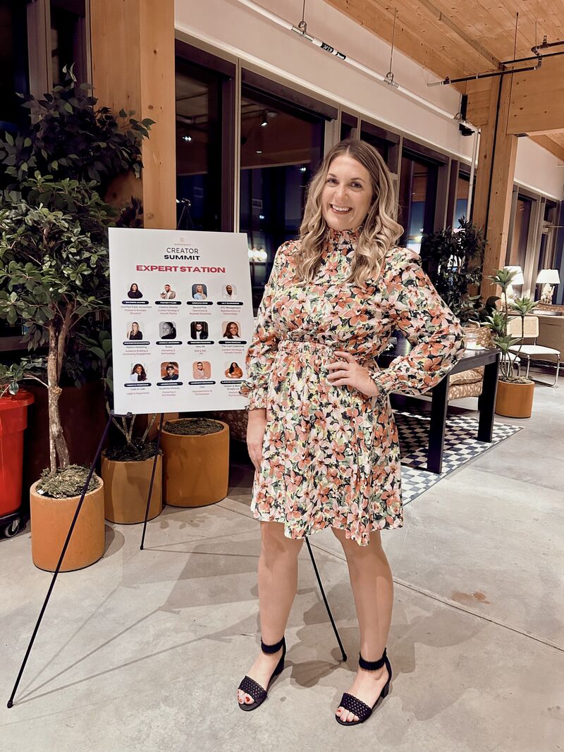 Woman in floral dress smiling indoors near a sign and plants, with a modern interior background.