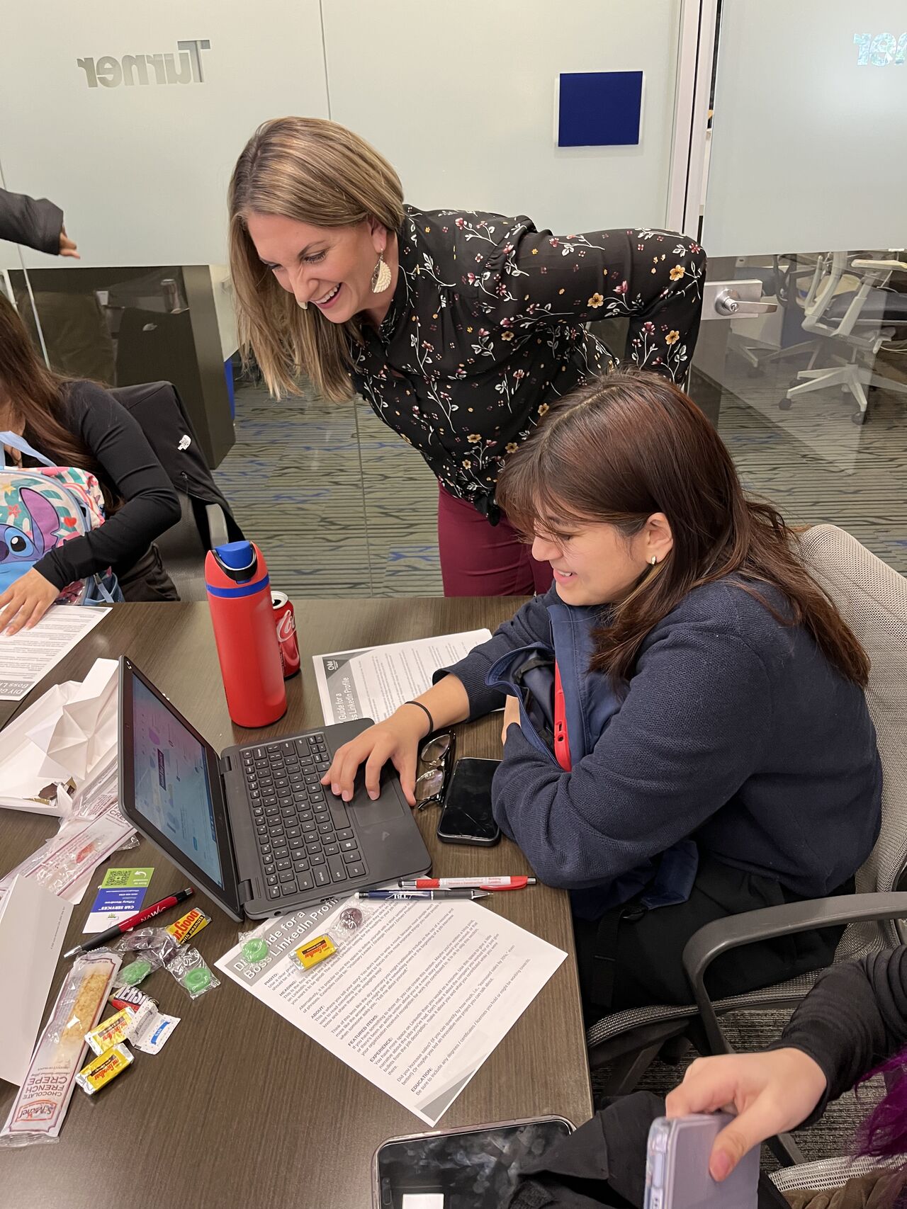 Two women in a meeting room, one standing and leaning over a seated woman working on a laptop, with papers and supplies on the table.