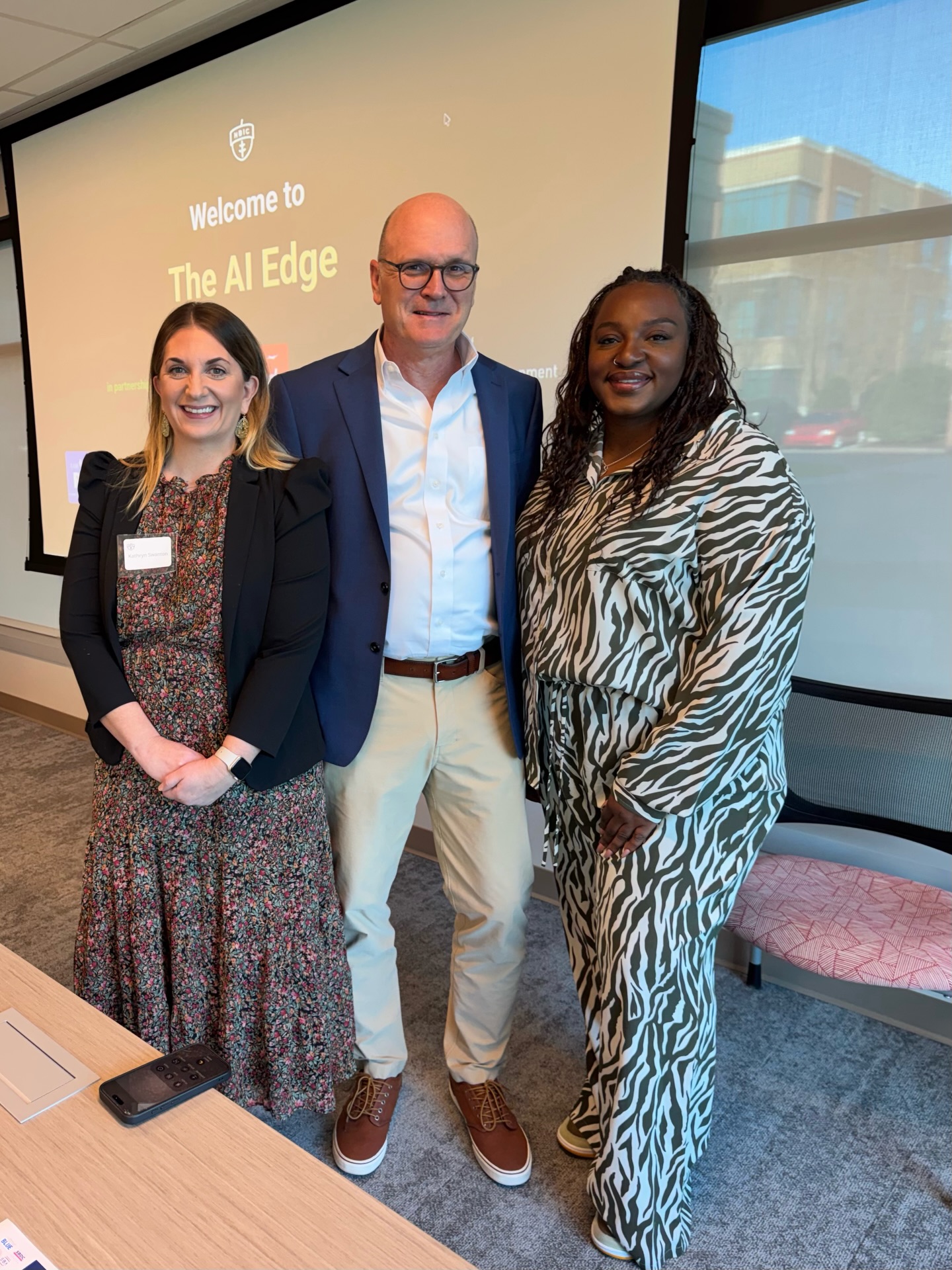 Three people standing indoors in front of a screen displaying 'Welcome to The AI Edge.' They are smiling and dressed professionally.