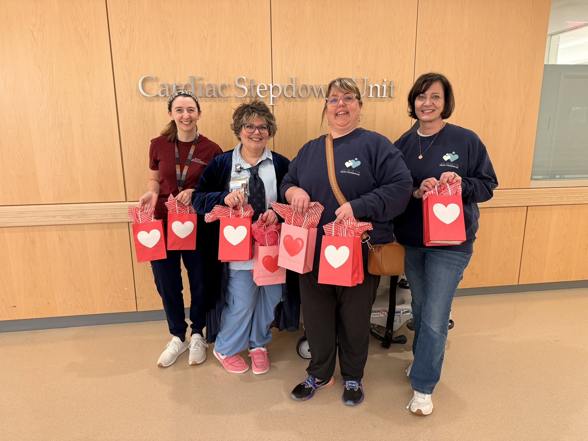 Four women stand in front of a wooden wall with a sign, holding gift bags with heart symbols, smiling.