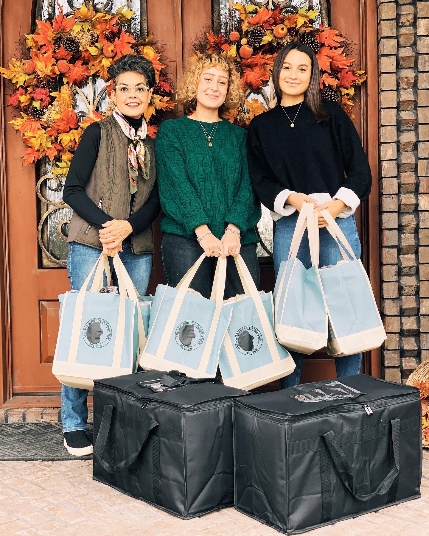 Three women stand in front of a decorated door with autumn flowers, holding gift bags, with large black bags on the ground.