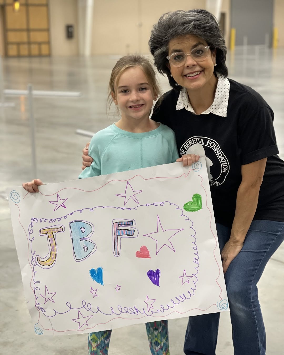 Two smiling females holding a colorful poster with stars and hearts, one adult and one child, in an indoor space.
