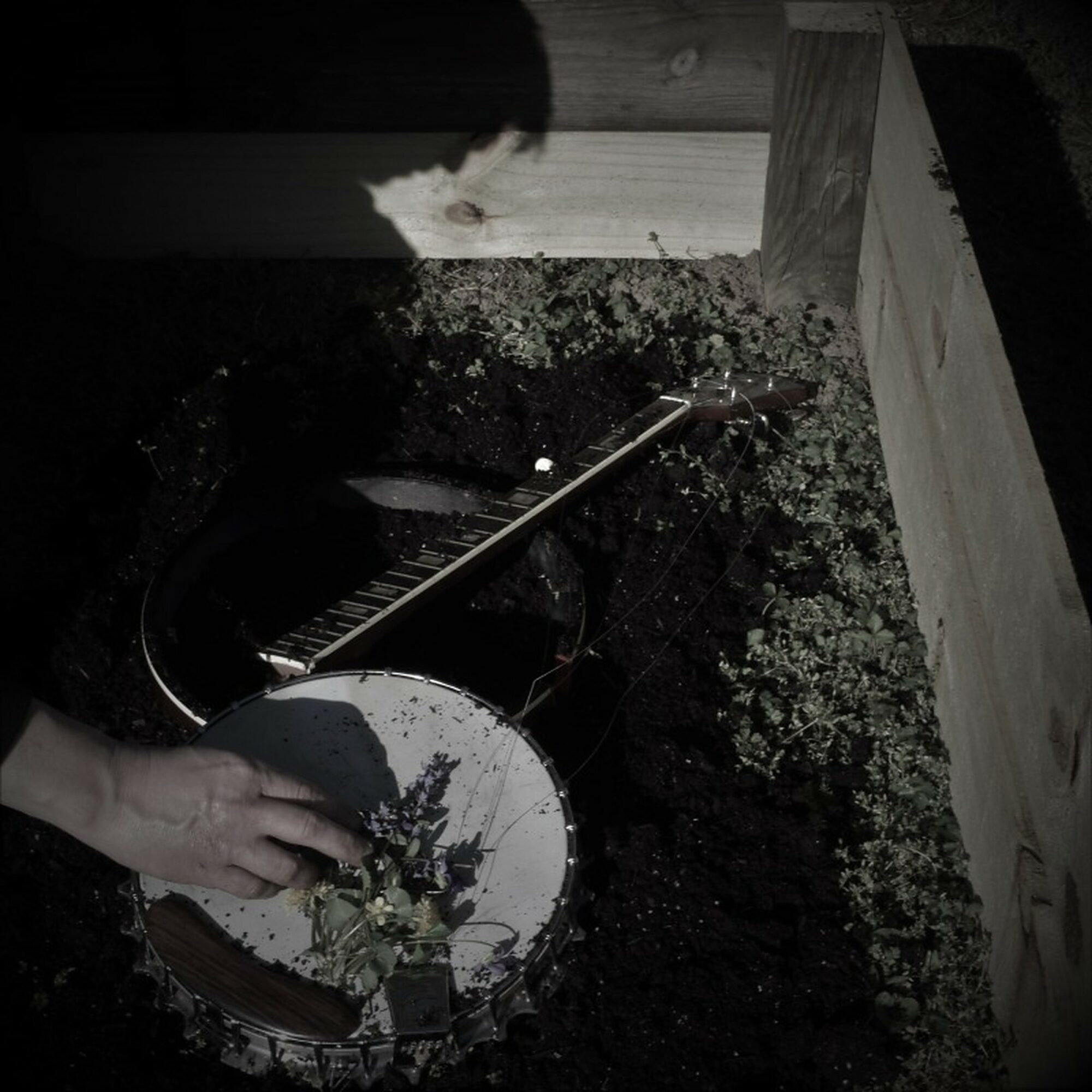 Person's hand playing a drum outdoors with a guitar nearby, surrounded by soil and wooden structure.