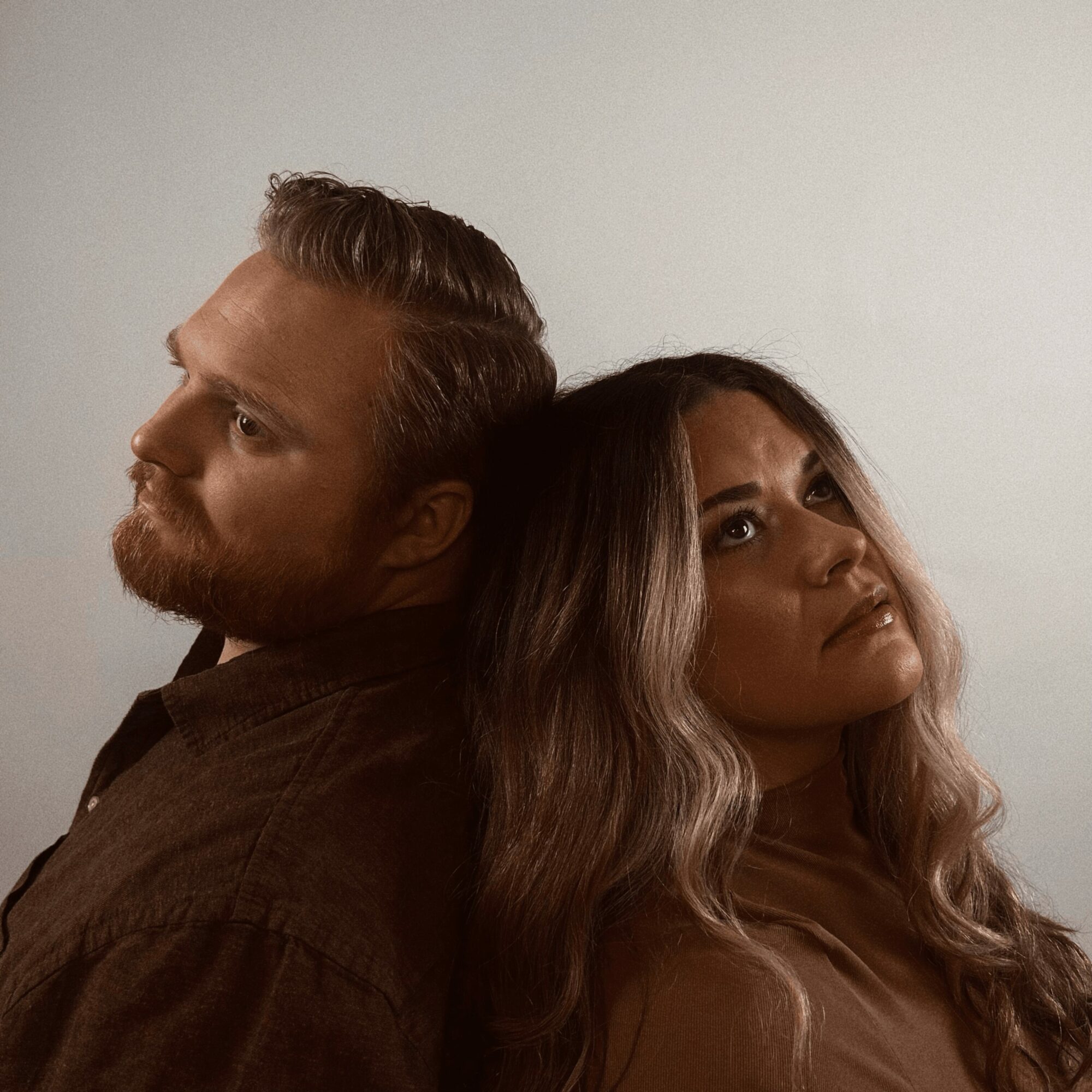 A man and woman are back-to-back, looking in opposite directions, with neutral expressions. The background is plain white.