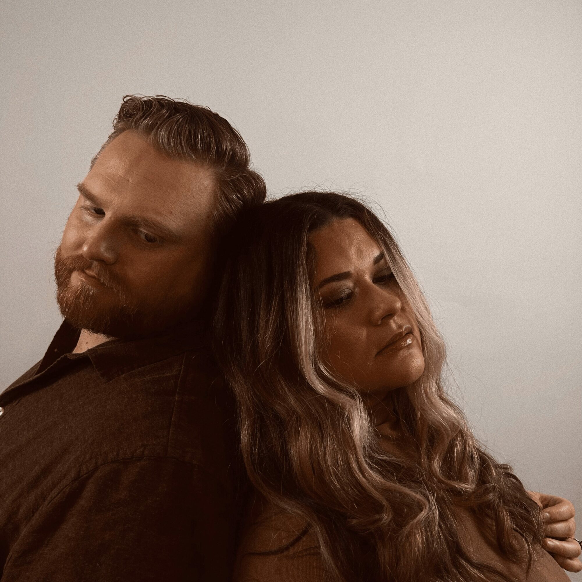 Two people with their heads close together, one with a beard and the other with long wavy hair, against a plain background.