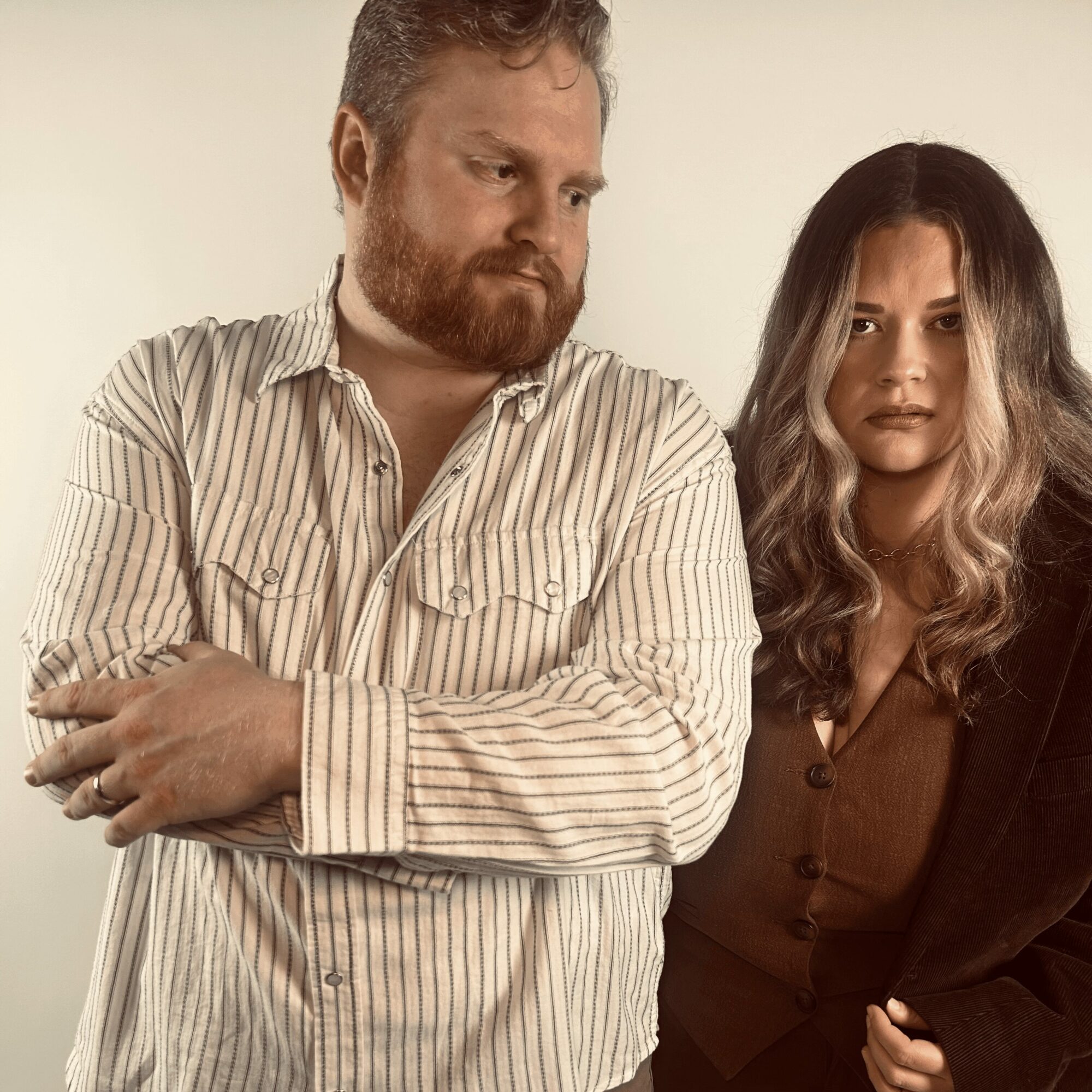 Two people standing close, one with arms crossed, both looking serious, against a plain background.