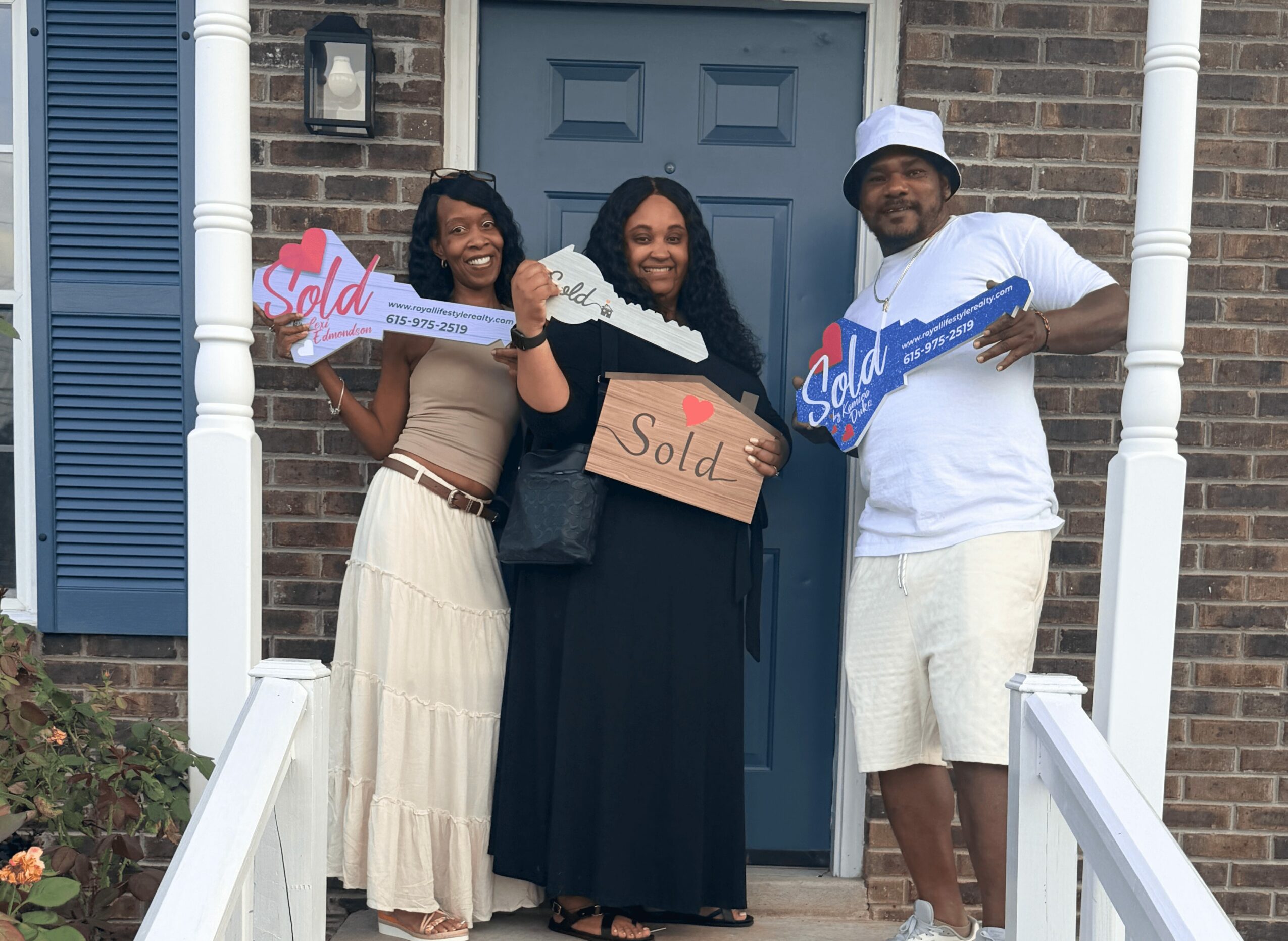Three people standing on front porch holding signs, smiling, in front of brick house with blue shutters.