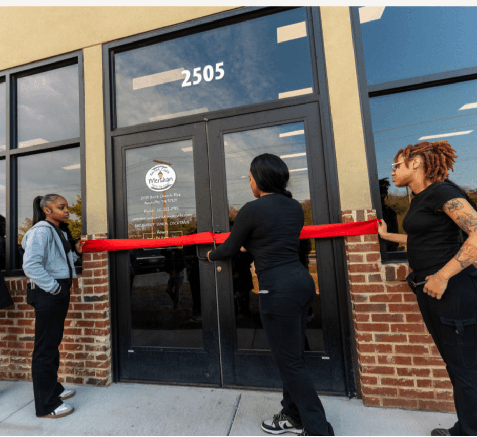 Three women participate in a ribbon-cutting ceremony outside a building with the number 2505, glass doors, and brick accents.