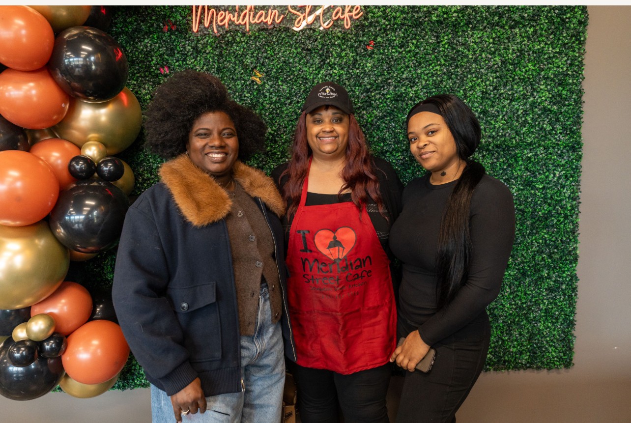 Three women standing in front of a green wall with balloons on the left side, smiling at the camera.