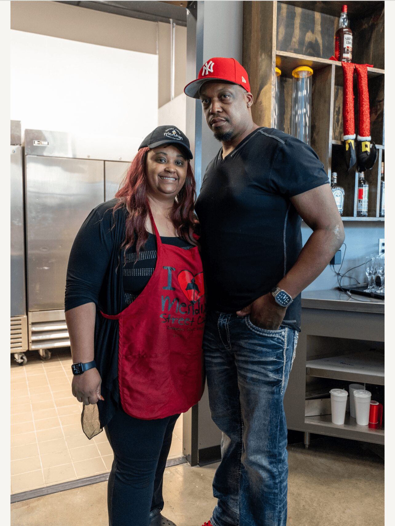 Two people standing indoors, one woman wearing a red apron and black cap, one man in a black shirt and red cap, smiling.