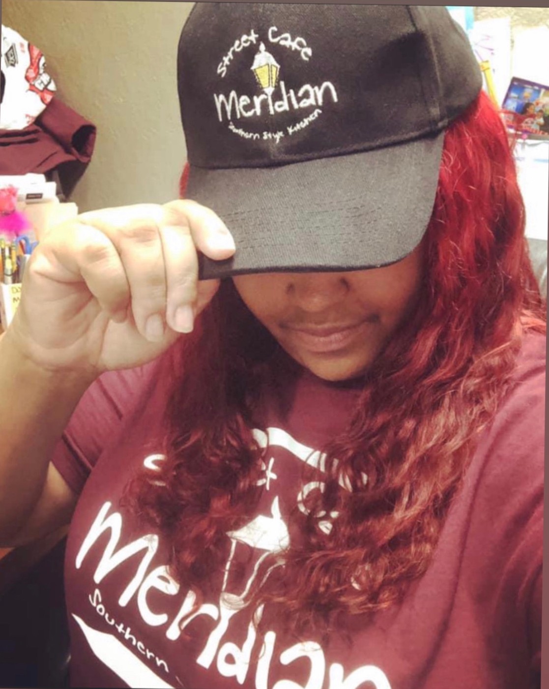 Person wearing a black cap with logo and text, holding the brim, and a maroon shirt with white text, curly hair visible.
