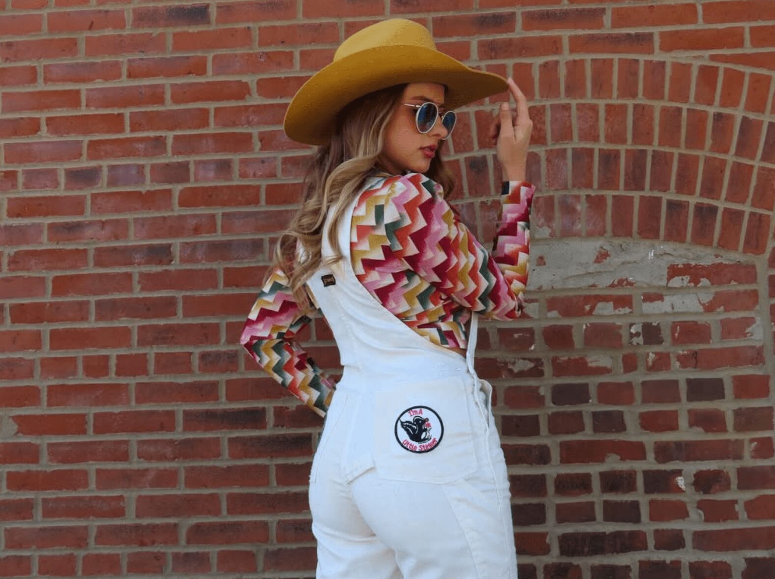Young woman wearing a wide-brimmed hat, sunglasses, colorful patterned top, and white overalls, standing against a brick wall.