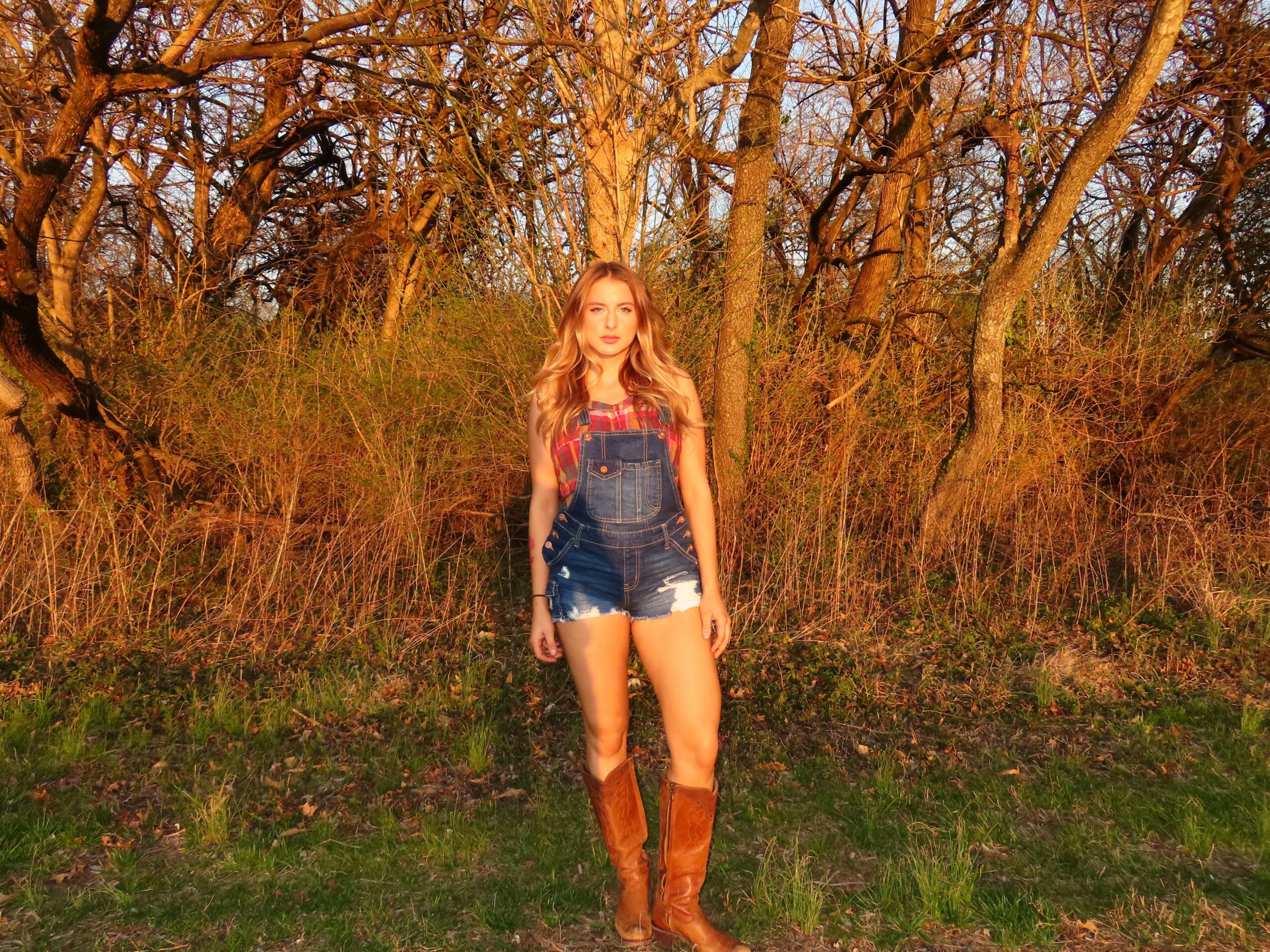 Young woman with long hair standing outdoors among trees and grass, wearing denim shorts and tall boots.