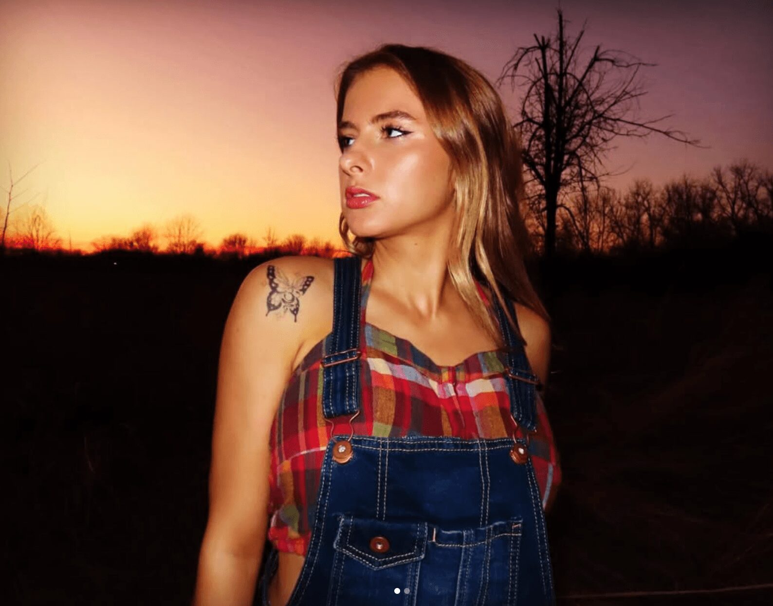 Young woman with long hair and a tattoo on her shoulder, wearing a plaid shirt and denim overalls, outdoors at sunset.