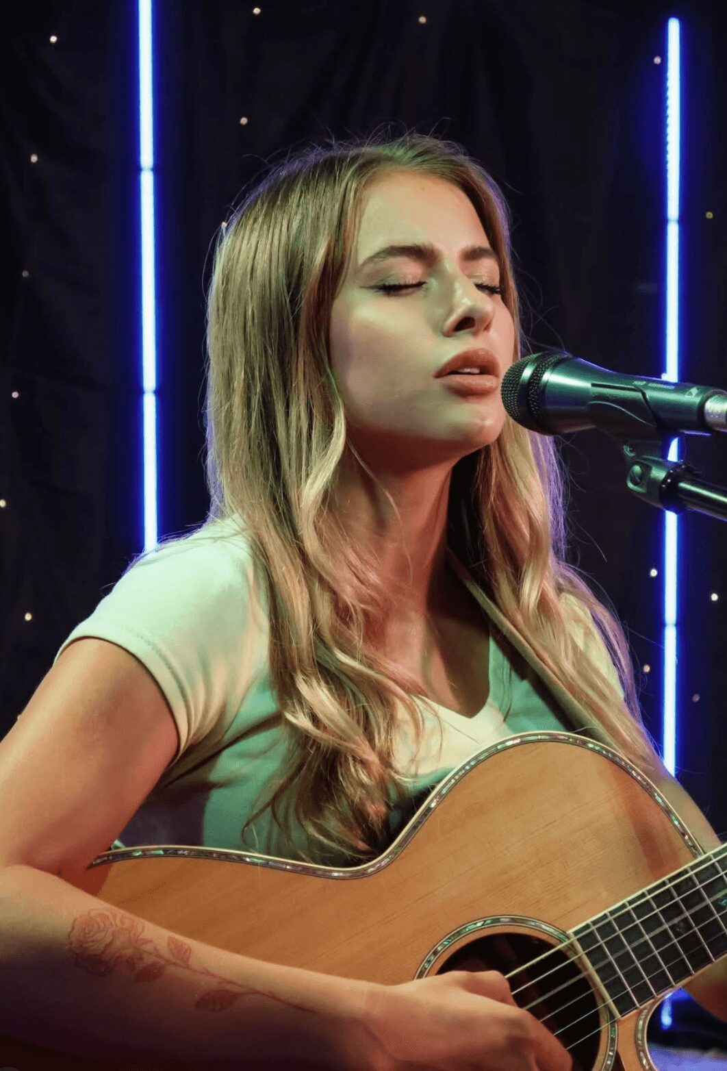 Young woman with long hair singing into a microphone while playing an acoustic guitar, with blue vertical lights behind her.