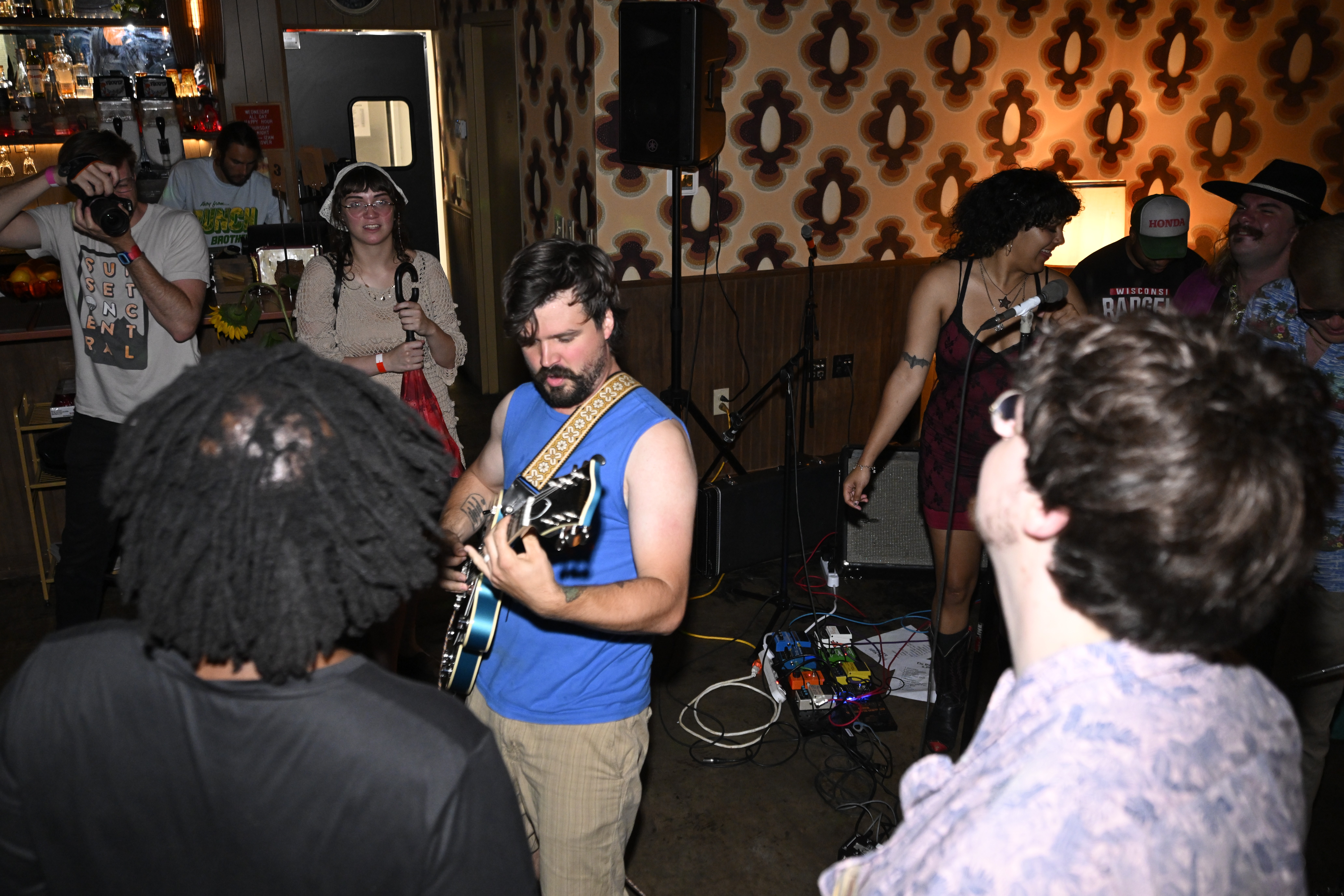 Group of people at a social gathering, some listening to a man playing guitar, others talking and dancing, in a room with patterned wallpaper.