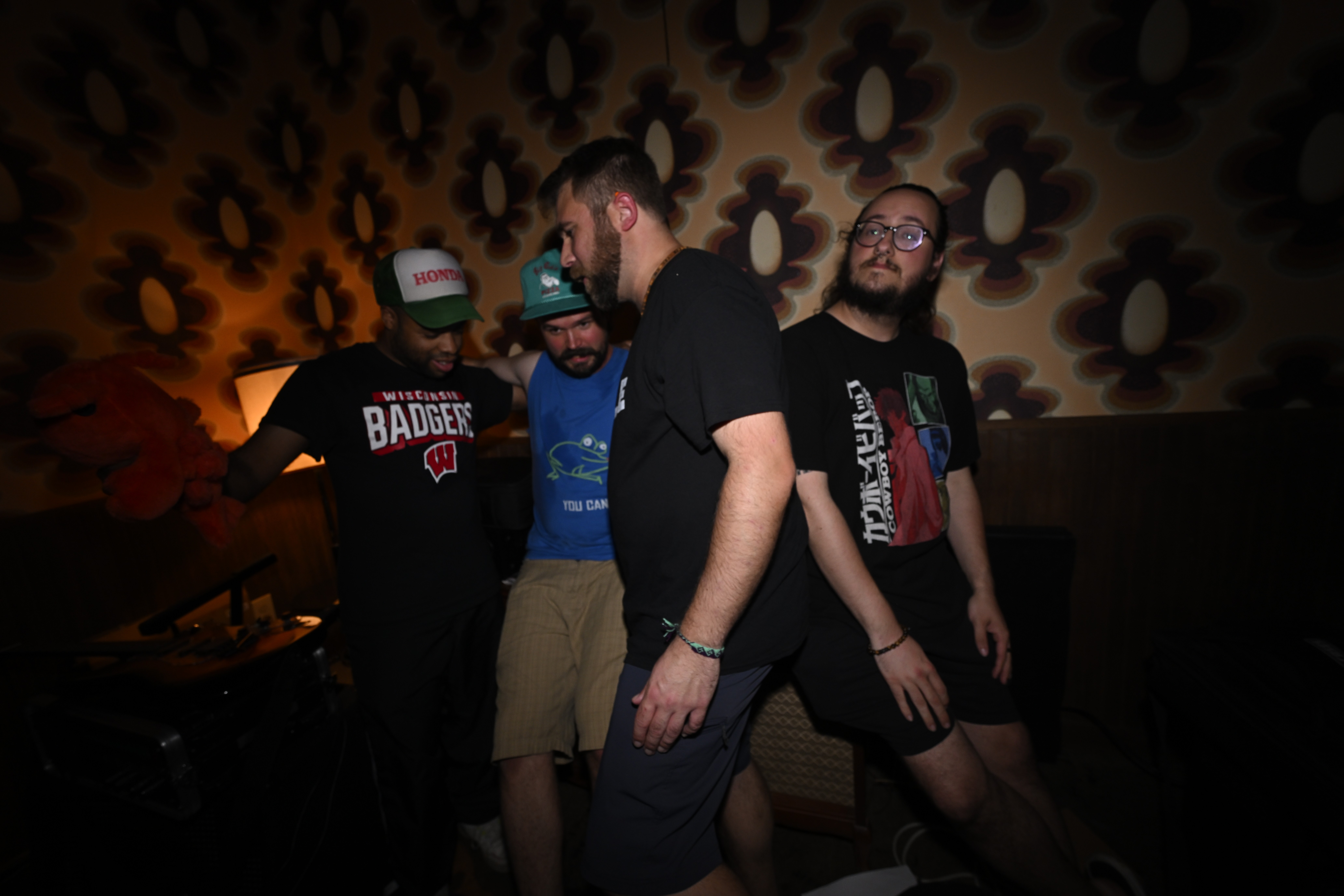 Four men standing together in a dimly lit room with floral wallpaper, some looking at the camera and others looking away.