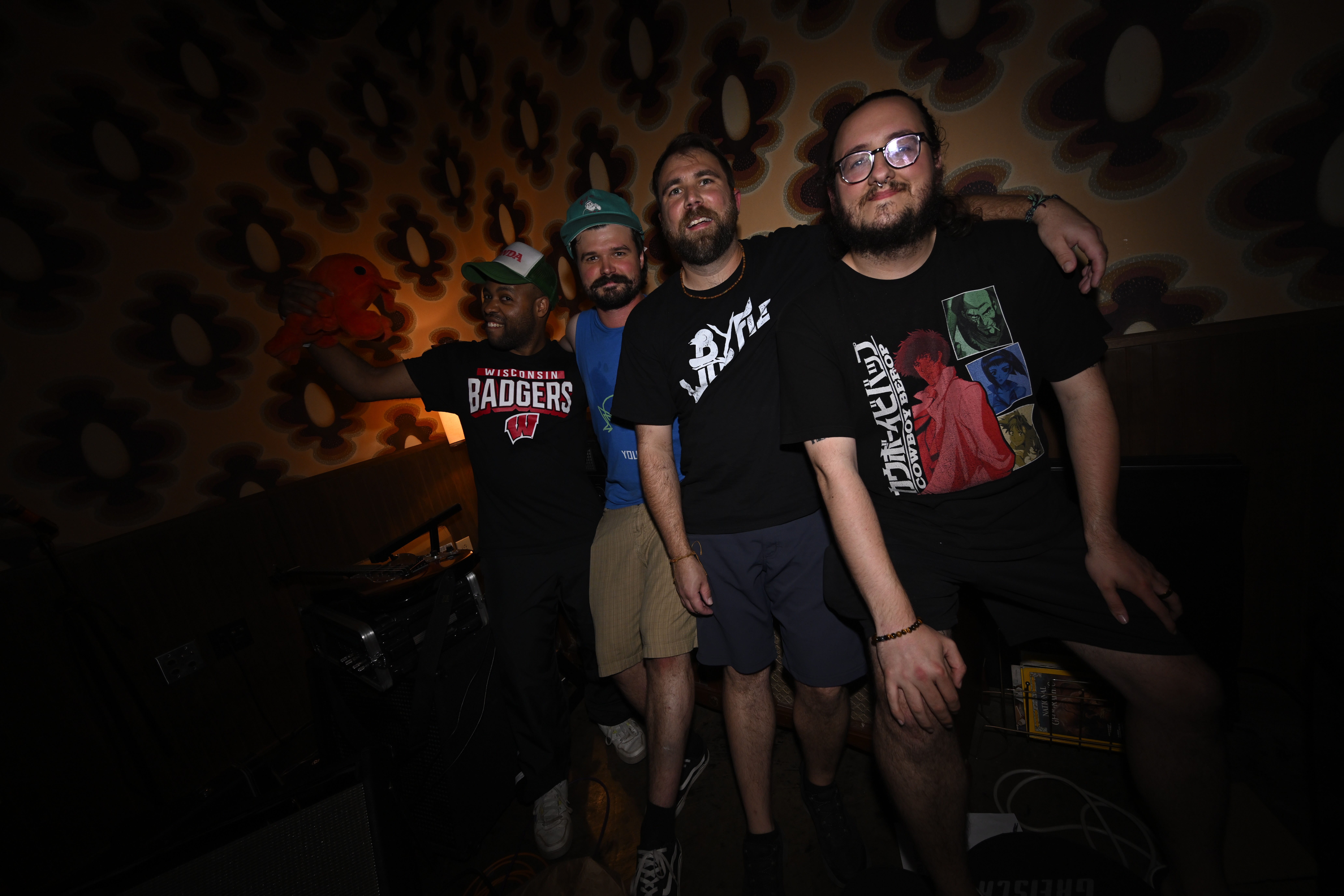 Four people standing together indoors with patterned wallpaper background, smiling and posing for the photo.