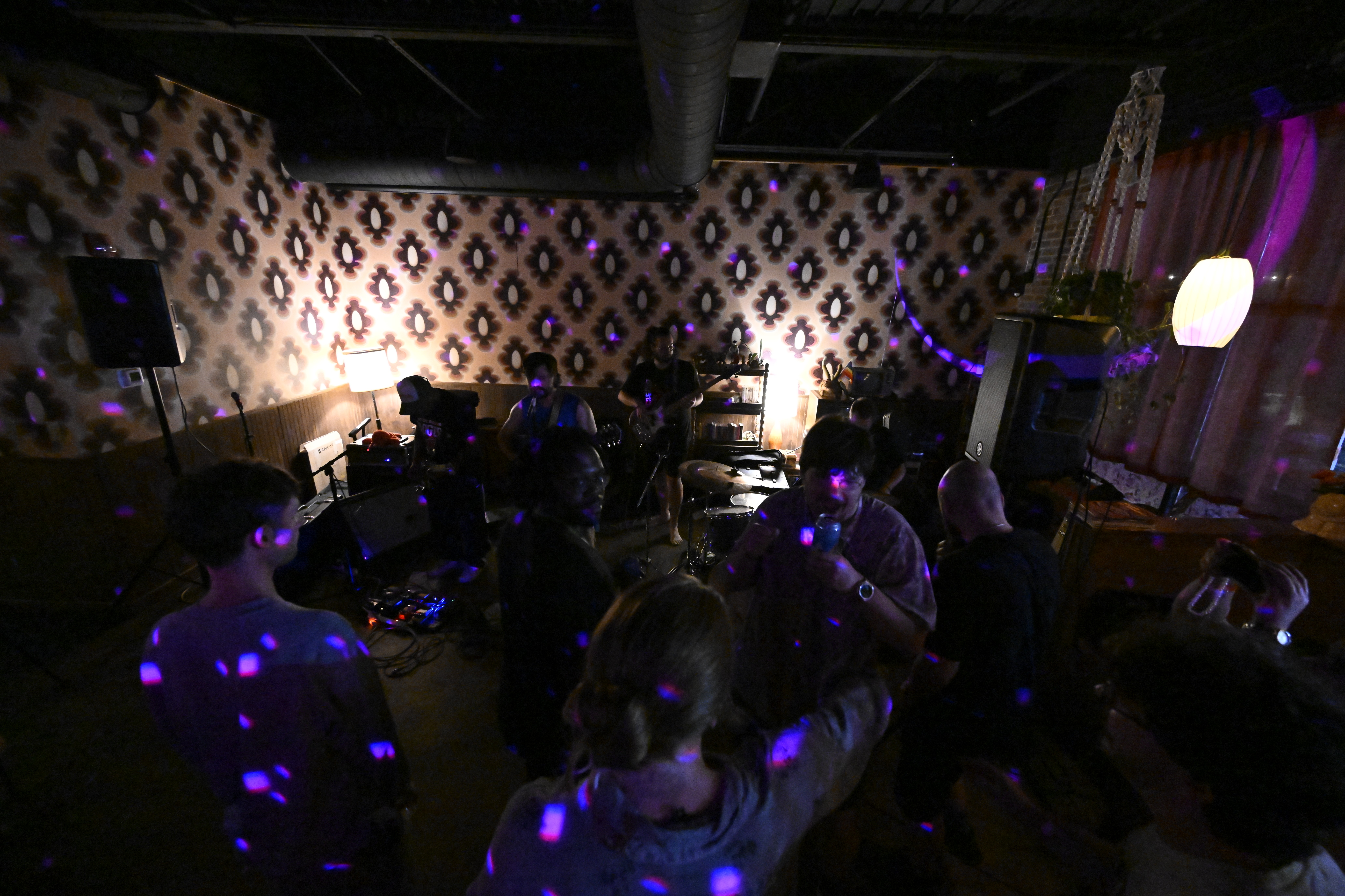 People gather in a dimly lit room with patterned wallpaper, illuminated by purple and white lights, some playing musical instruments.