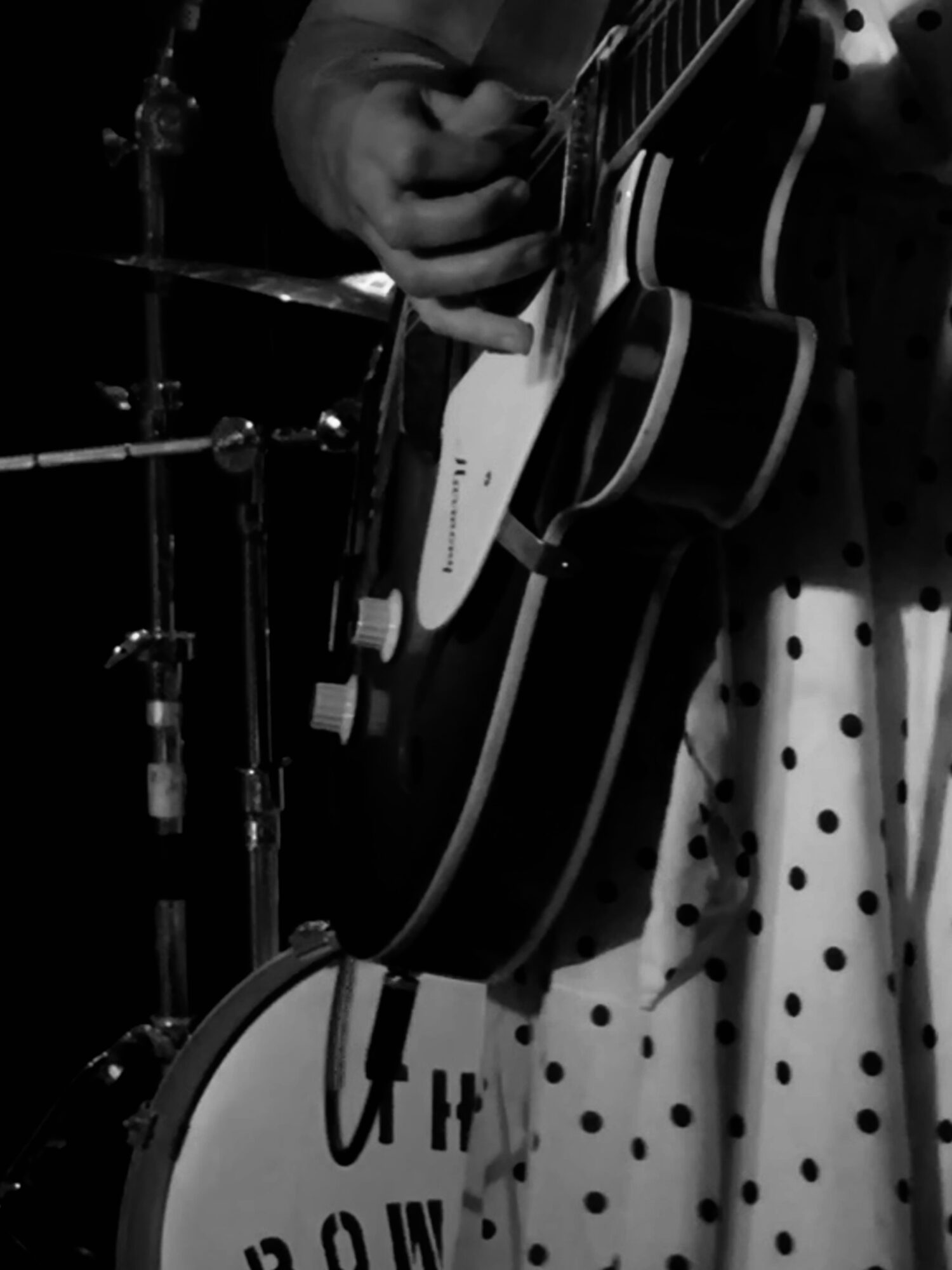 Person playing an acoustic guitar, wearing a polka dot dress, with a drum set in the background.