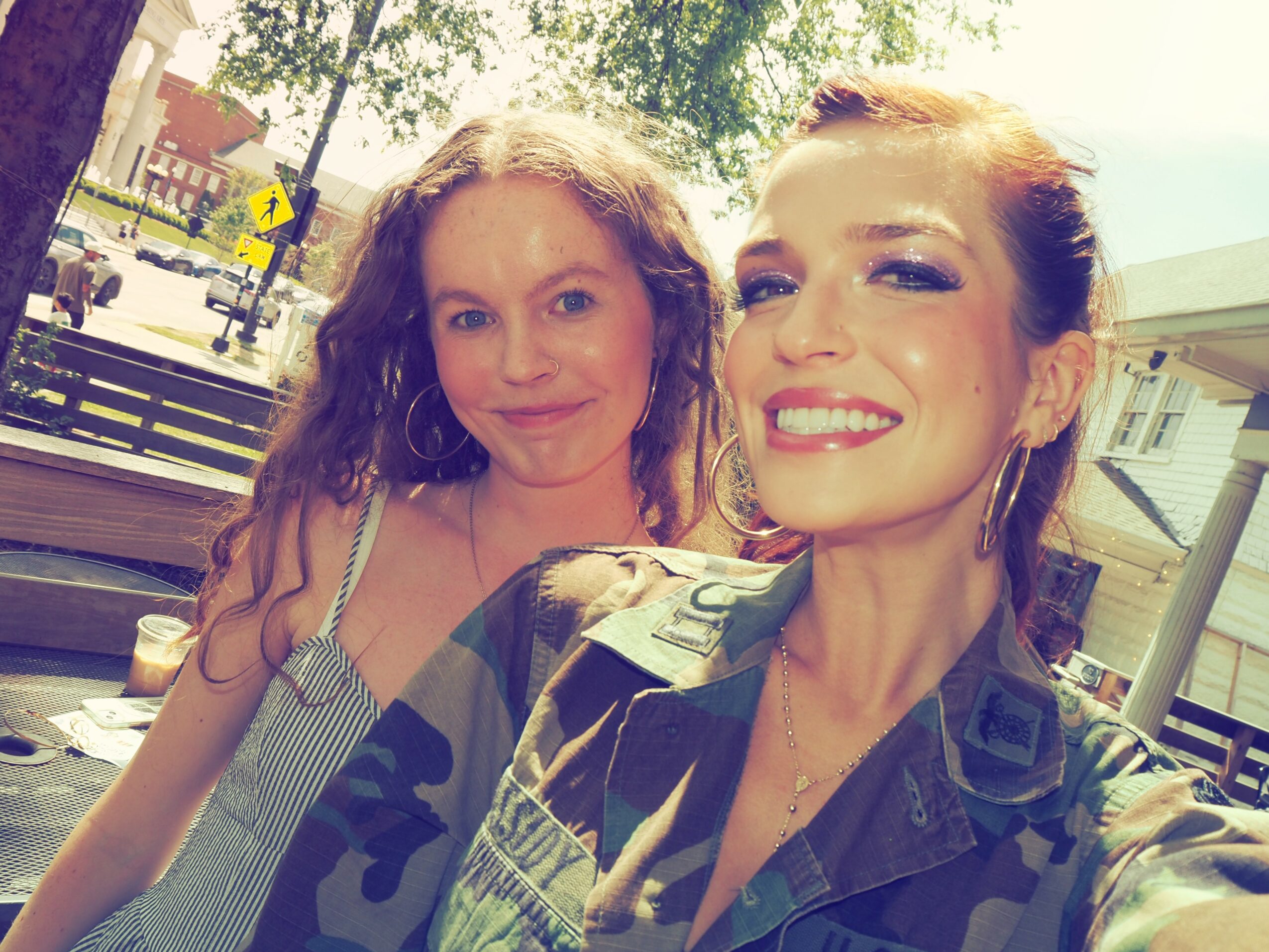 Two women smiling outdoors, one with curly hair and the other with straight hair, in bright sunlight.