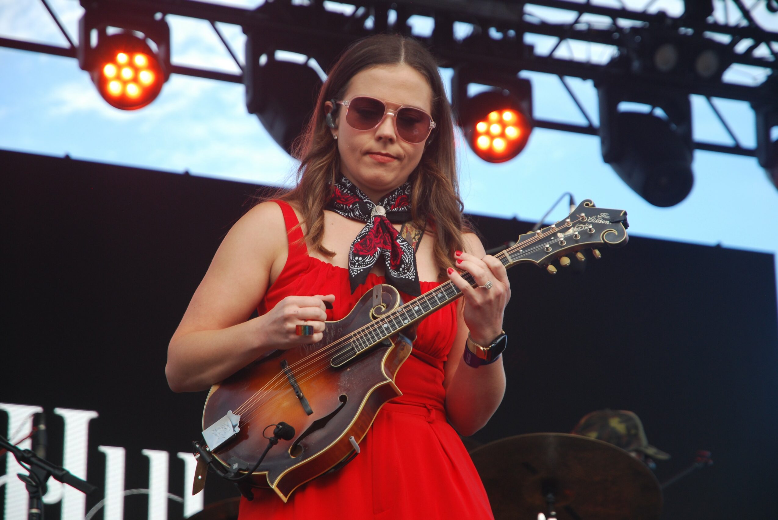 Woman in red dress playing a string instrument on stage with lights and a black background.