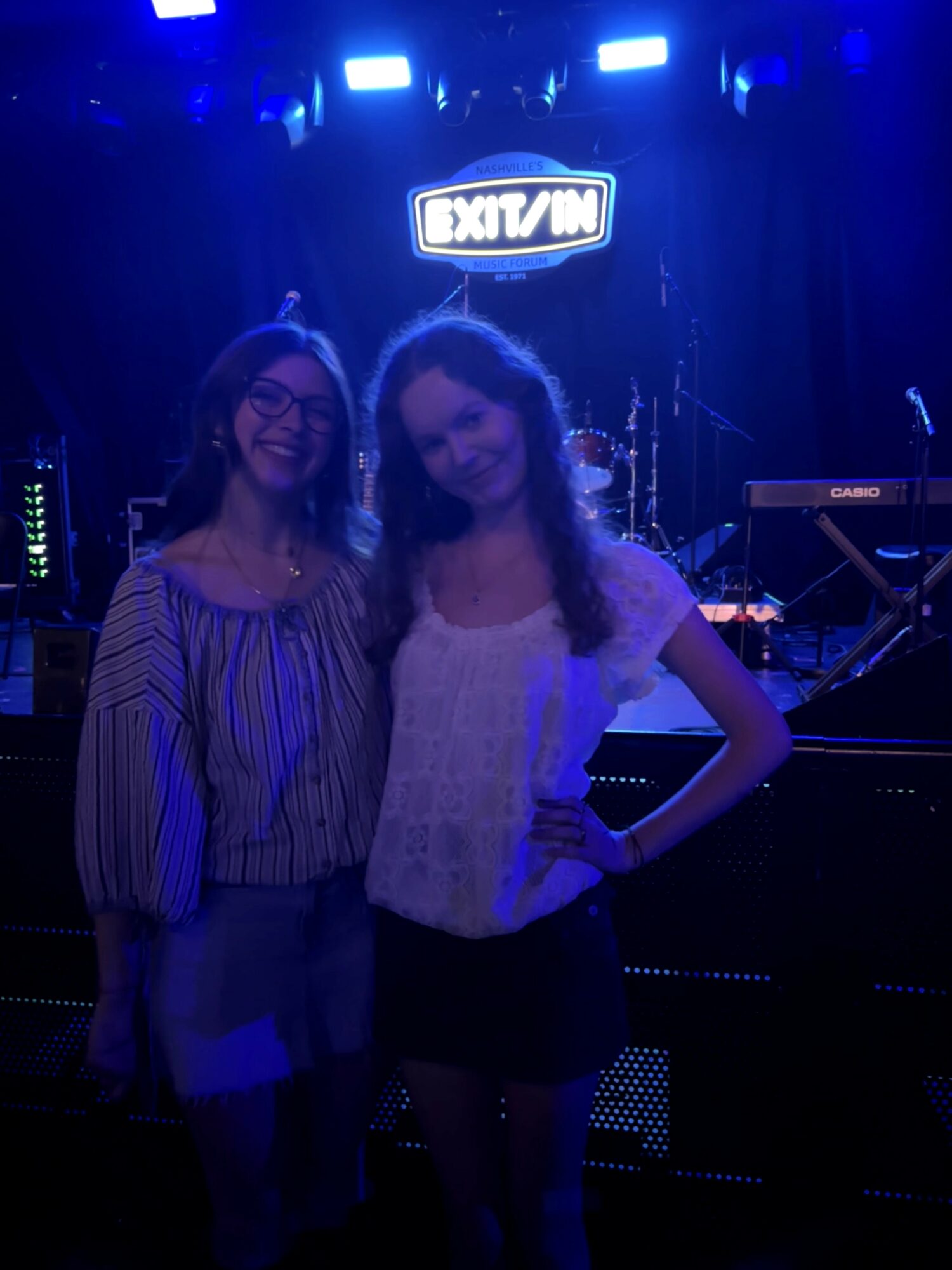 Two women stand on a stage with musical instruments and a sign that reads 'EXIT/IN' behind them.