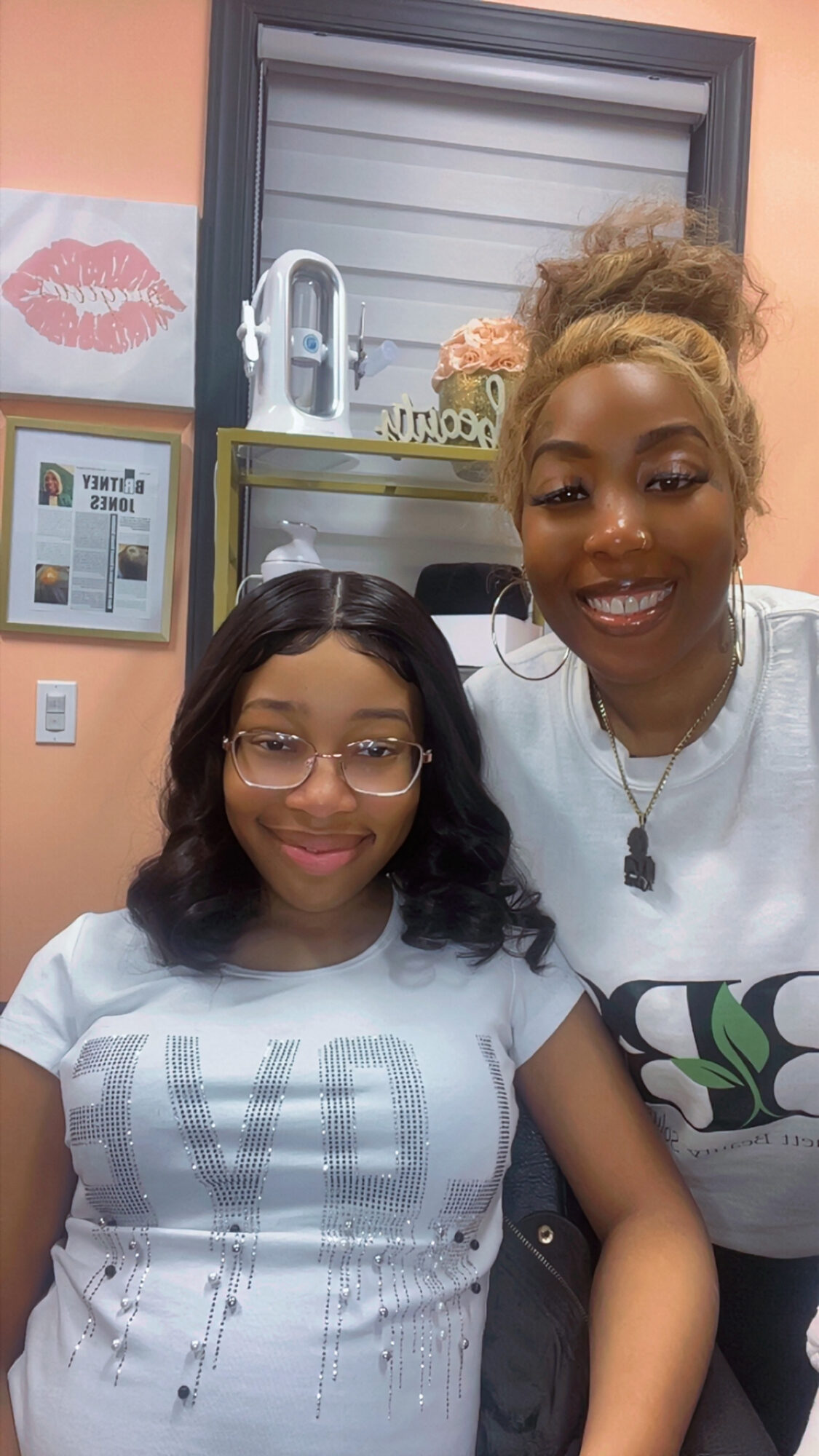 Two smiling women pose indoors, one with glasses and curly hair, the other with blonde hair in a bun, background includes a window and framed pictures.