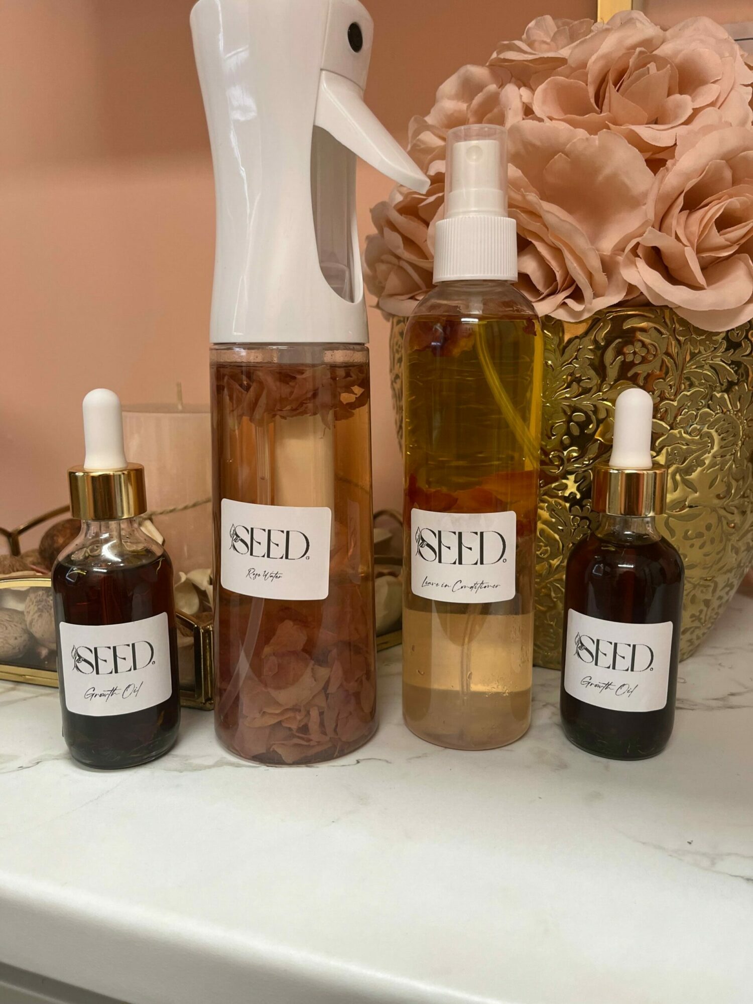 Four bottles with dropper caps and labels, arranged on a surface with a pink flower and decorative items in the background.