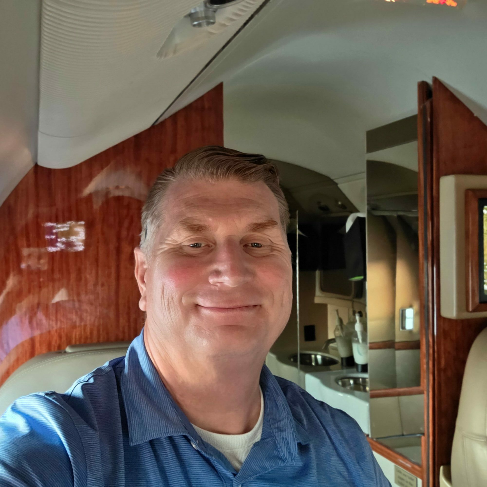 Person with short hair and a blue shirt sitting in an airplane seat, smiling, with a wooden panel and window behind.
