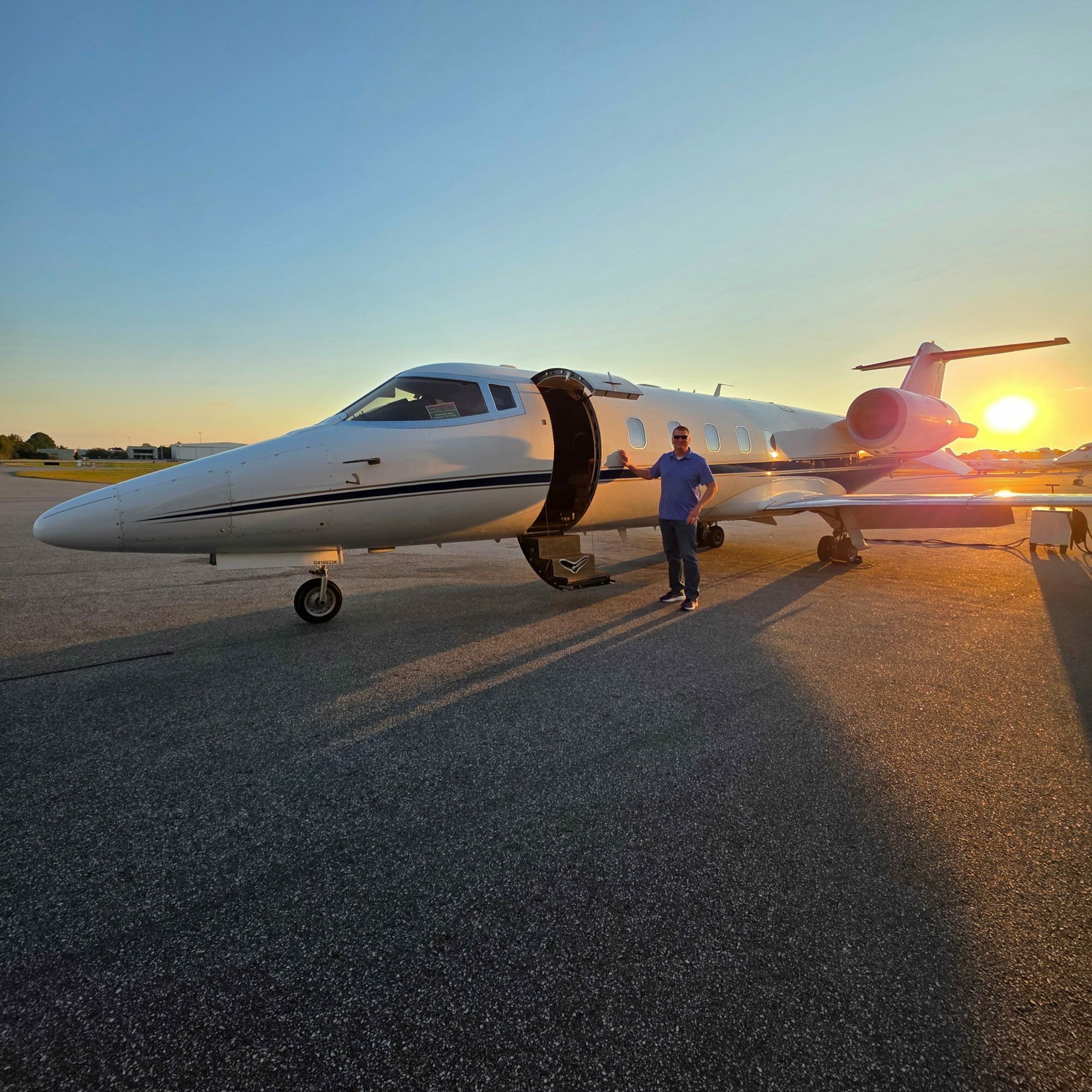 Person walking beside a parked airplane on a runway during sunset, with the sun low in the sky.