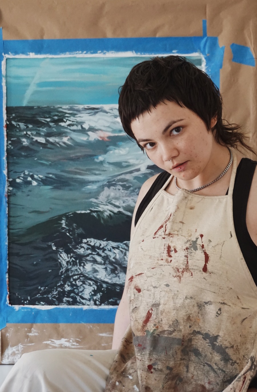 Person with short dark hair wearing a stained apron and backpack, standing in front of a painting of water, looking at the camera.
