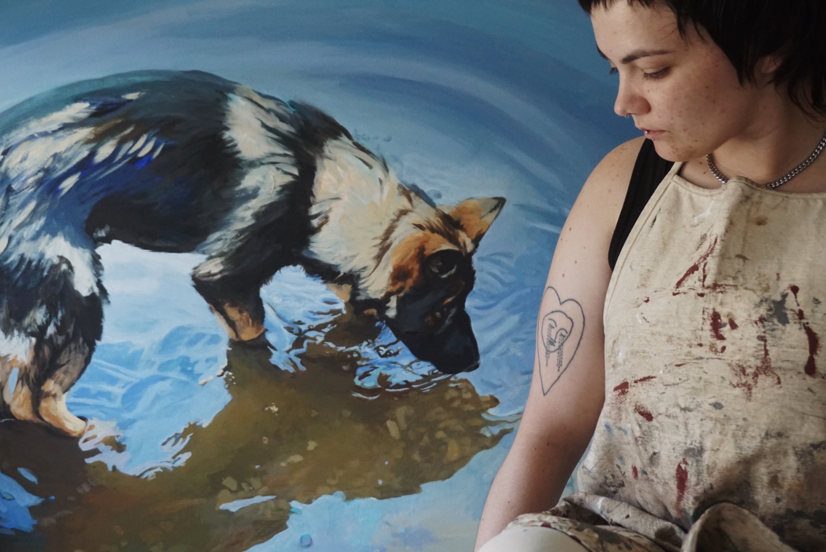 Person with short dark hair and a tattoo on arm looks at a dog in water, which is drinking from a puddle.