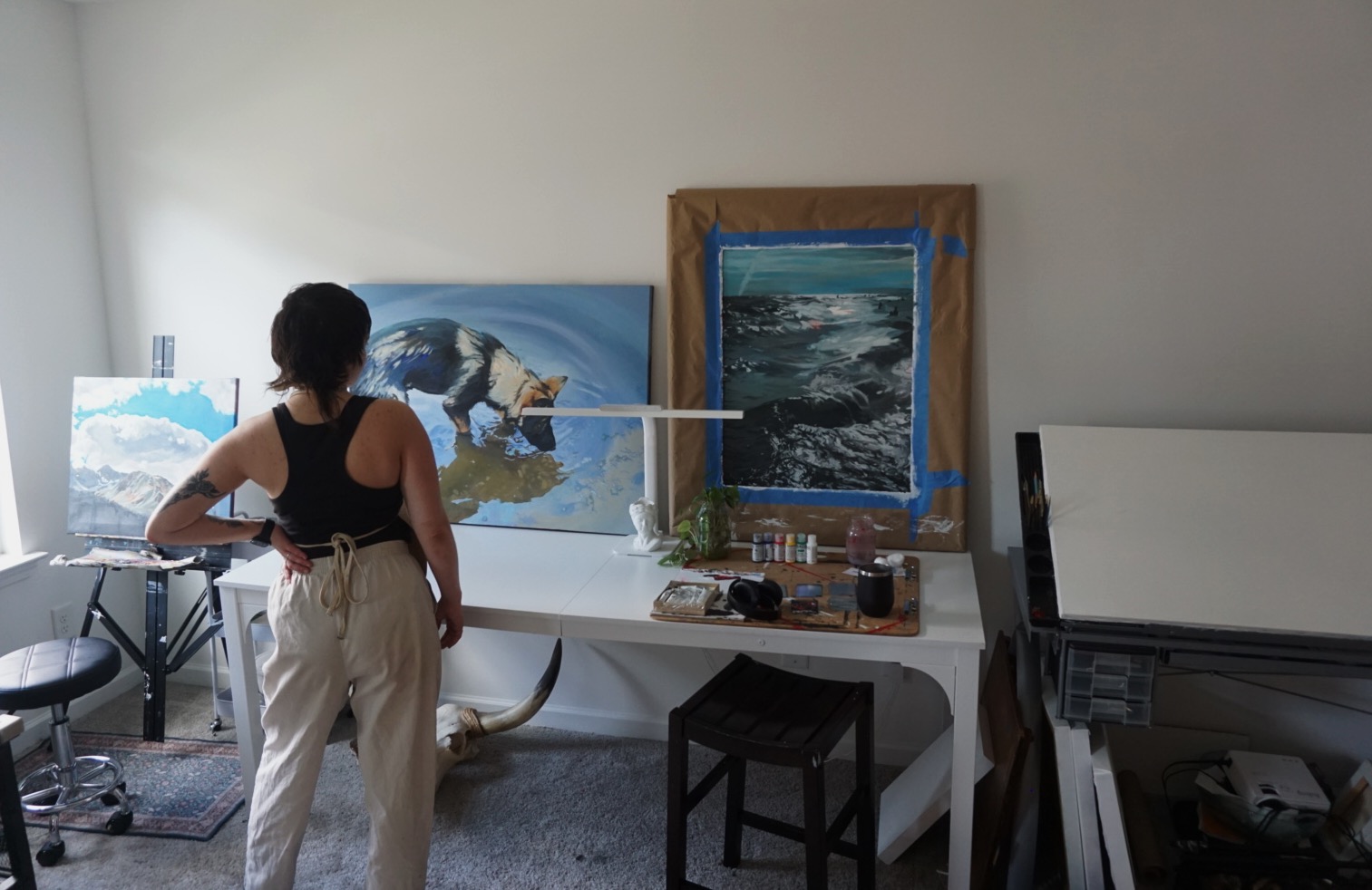 Person standing in art studio, painting ocean scene on canvas, with framed ocean picture and art supplies nearby.