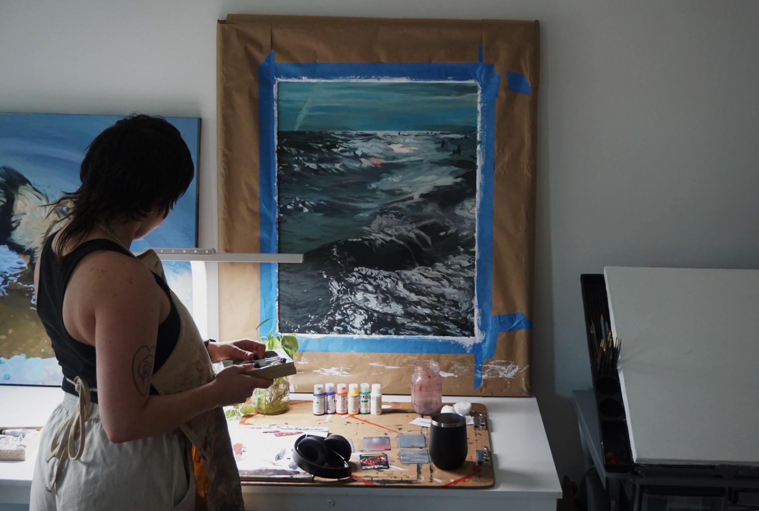 Artist painting a seascape with ocean waves, standing in front of a window with painting supplies on a table.