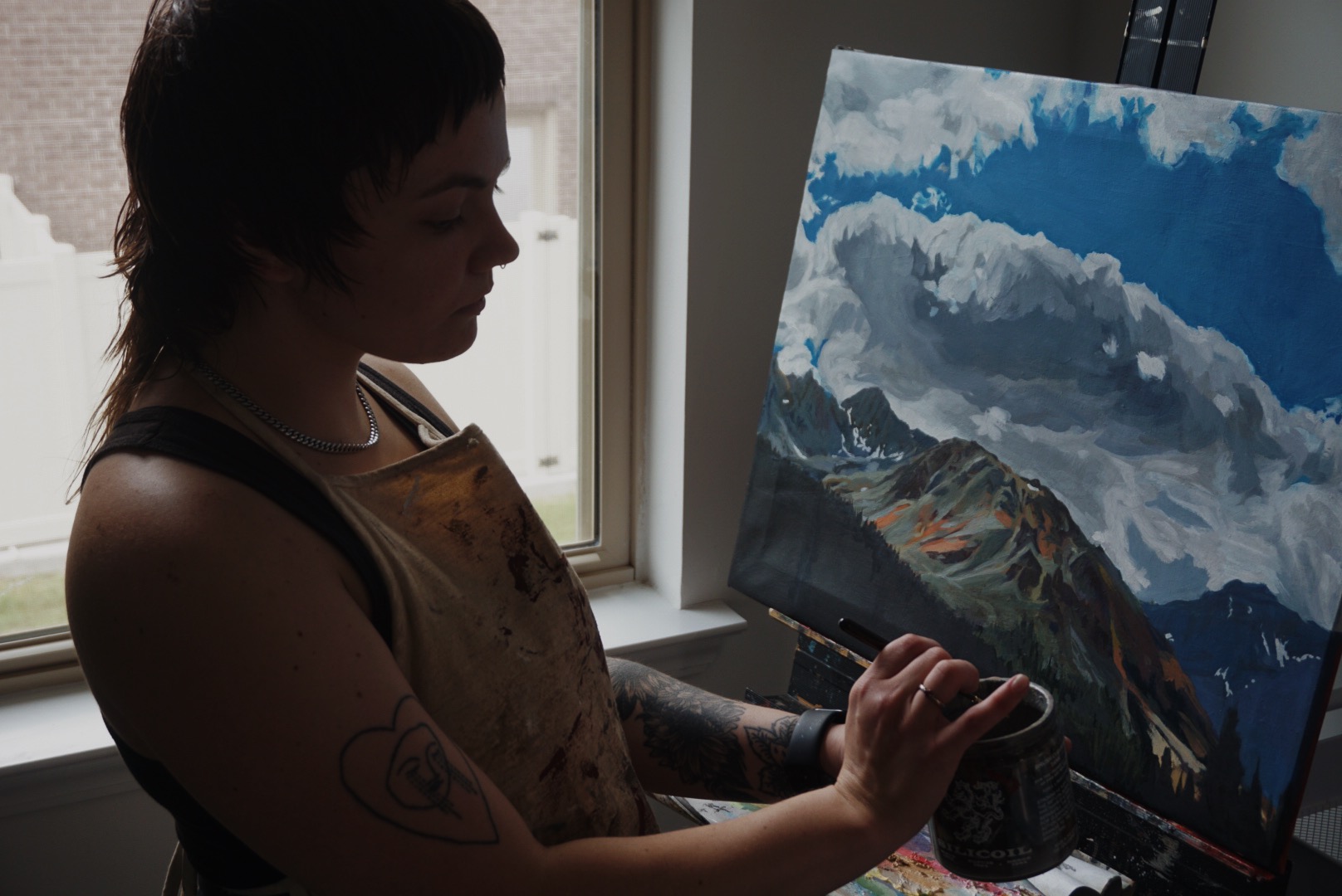Person with short dark hair painting on a landscape canvas near a window, holding a brush, with natural light illuminating the scene.