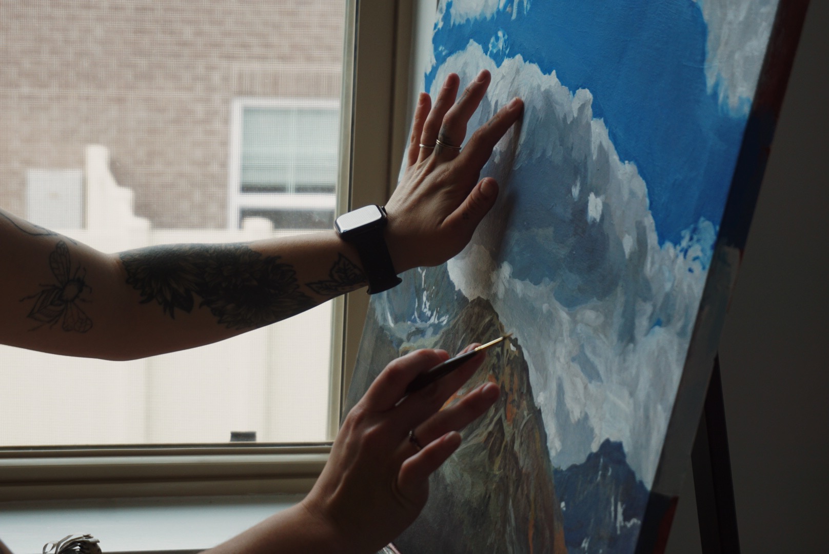 Person's hands touch a painting of mountains and sky on a digital screen, with a window in the background.