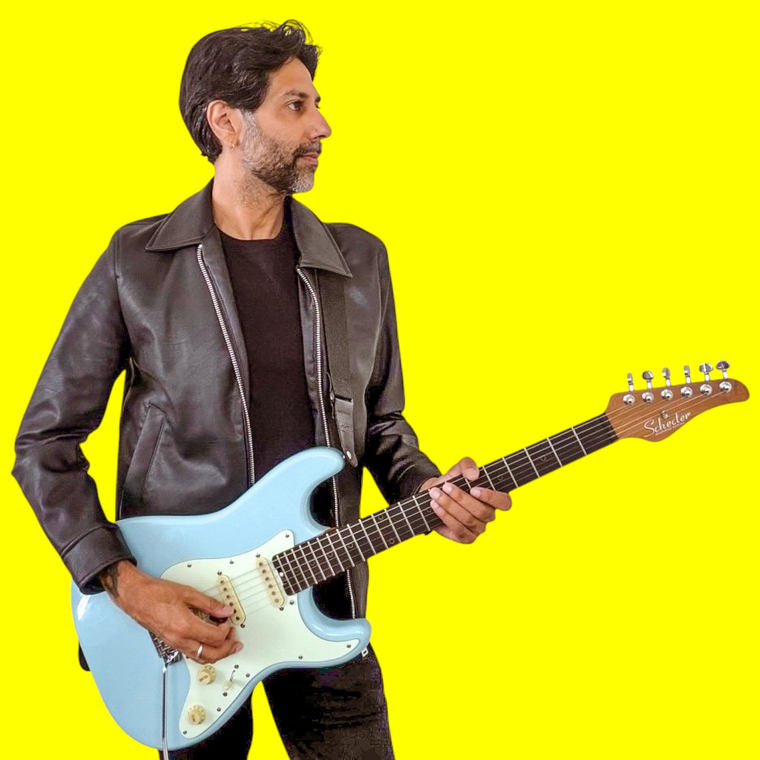 Man with dark hair and beard wearing a black leather jacket playing a light blue electric guitar against a yellow background.