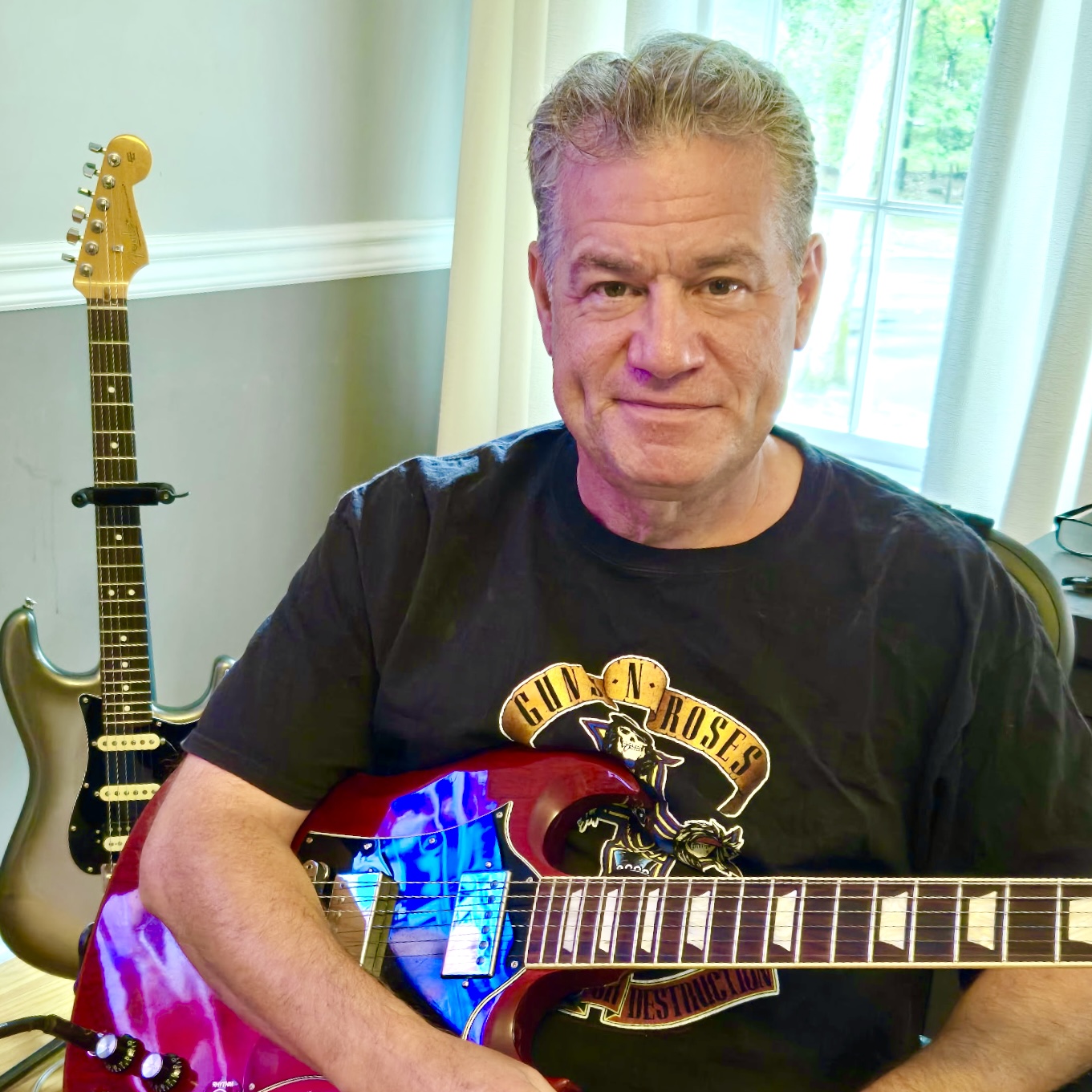Man sitting with guitar, electric guitar in background, wearing a black Guns N' Roses t-shirt, indoors near window.