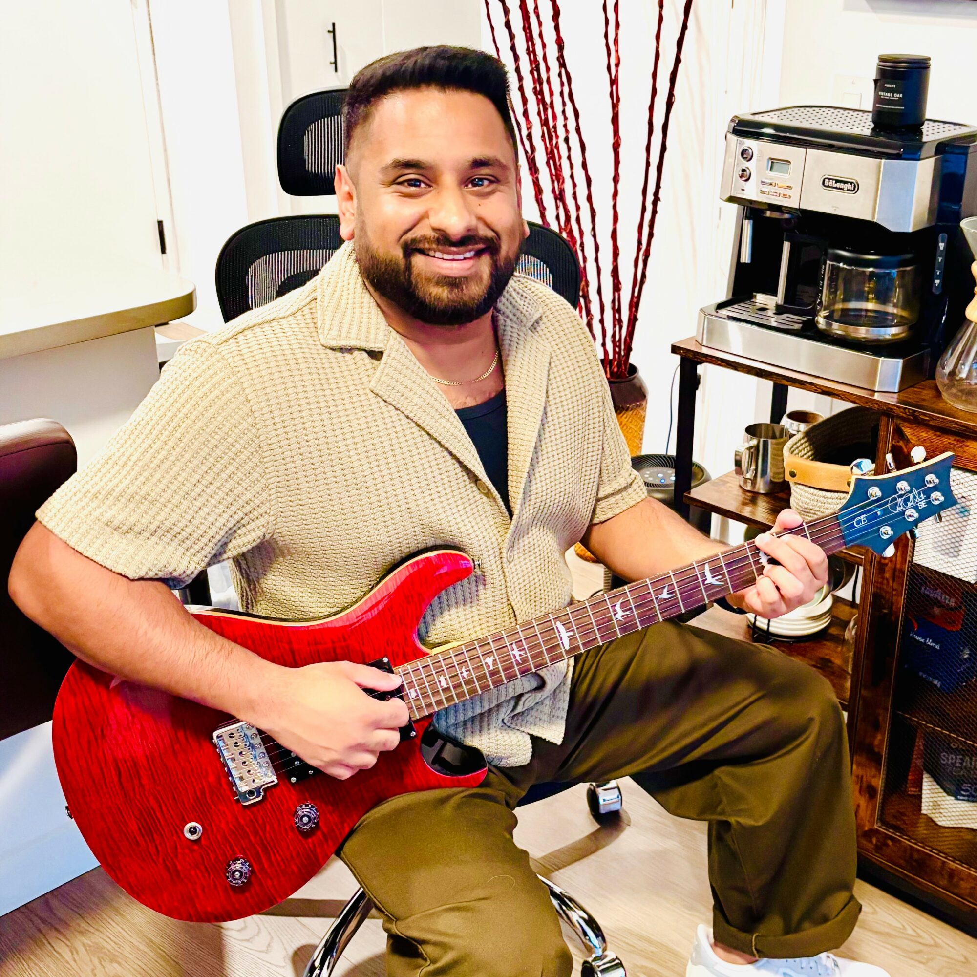 Man smiling, sitting on a chair, holding a red electric guitar in a room with kitchen appliances and decor.