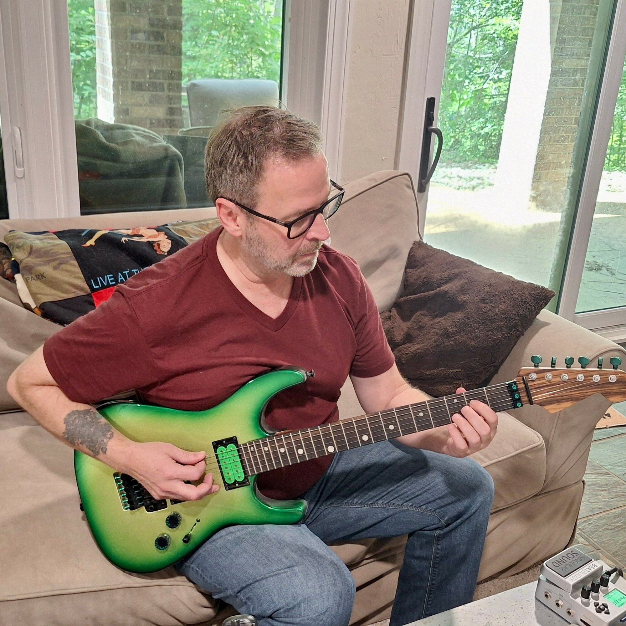 Man with glasses and a beard playing a green electric guitar on a beige sofa in a bright room.