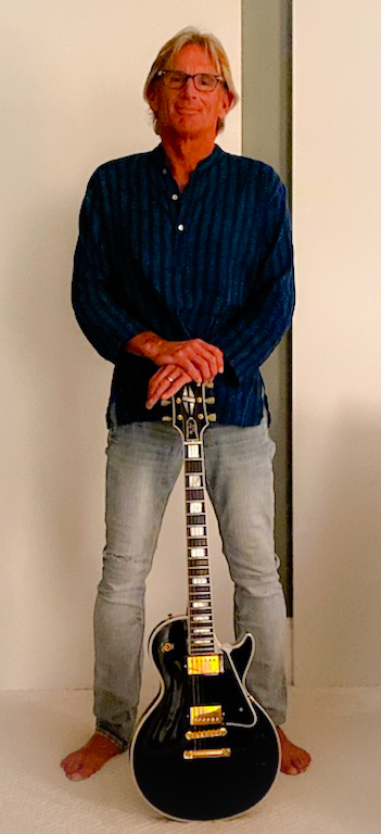 Person standing with a guitar, wearing glasses, a dark striped shirt, and light jeans, against a plain background.