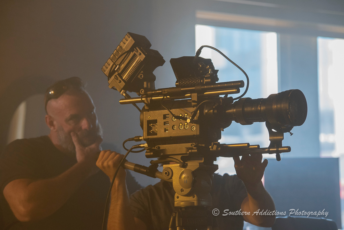 A man operating a professional video camera indoors near a window with sunlight, holding the camera steady.