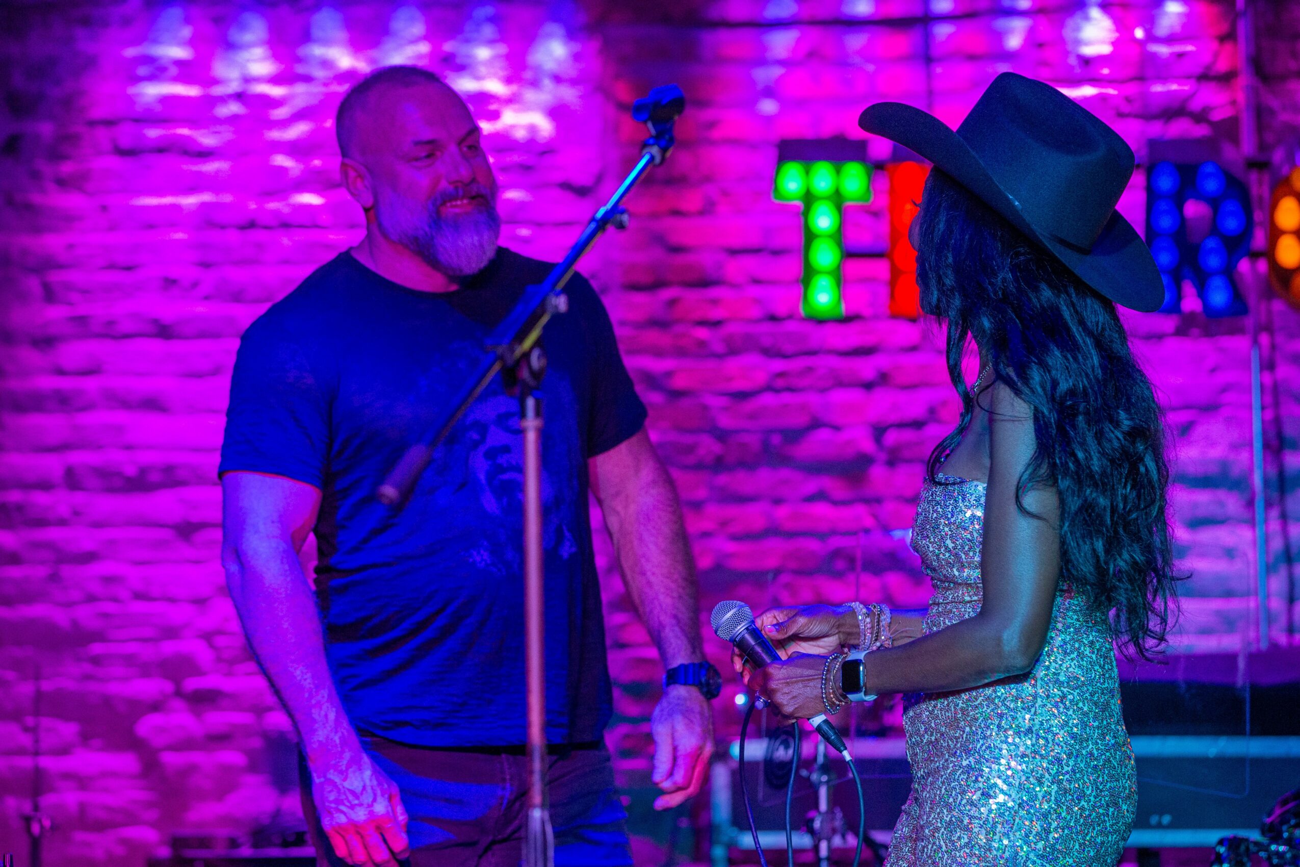 A man and woman on stage with a brick wall background, colorful lights, and a microphone stand, engaging in conversation.
