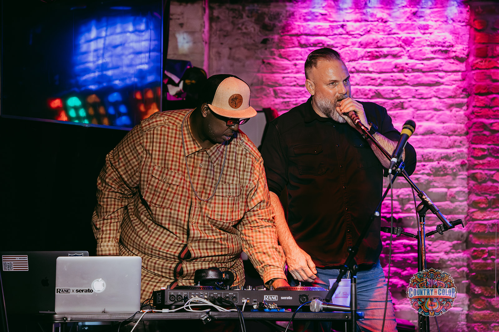 Two men perform on stage with a brick wall background, one DJ and one singer, colorful lighting, and equipment.