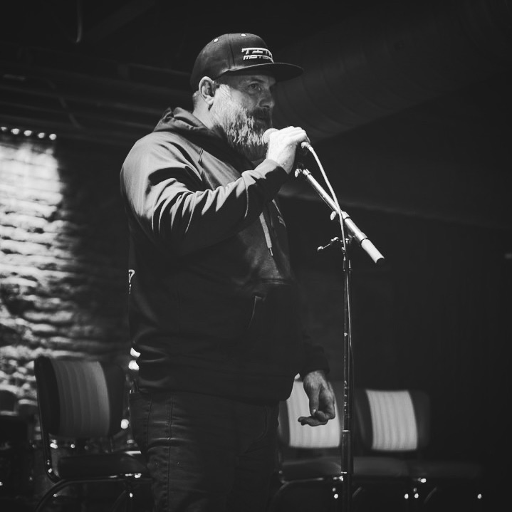 Man with beard and cap speaking into microphone on stage, with chairs and brick wall background, in black and white.