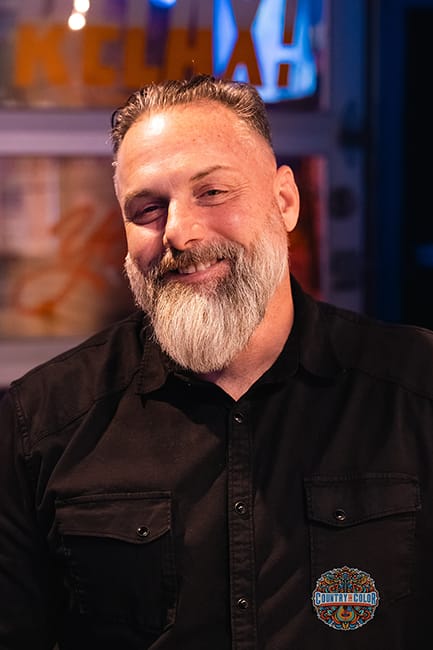 Man with a beard smiling, wearing a black shirt, in a colorful indoor setting.