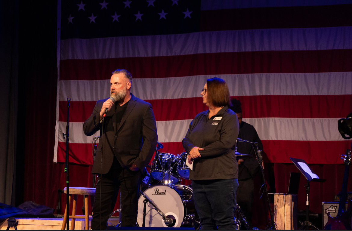 Two people stand on stage in front of an American flag backdrop, one speaking into a microphone, the other listening.