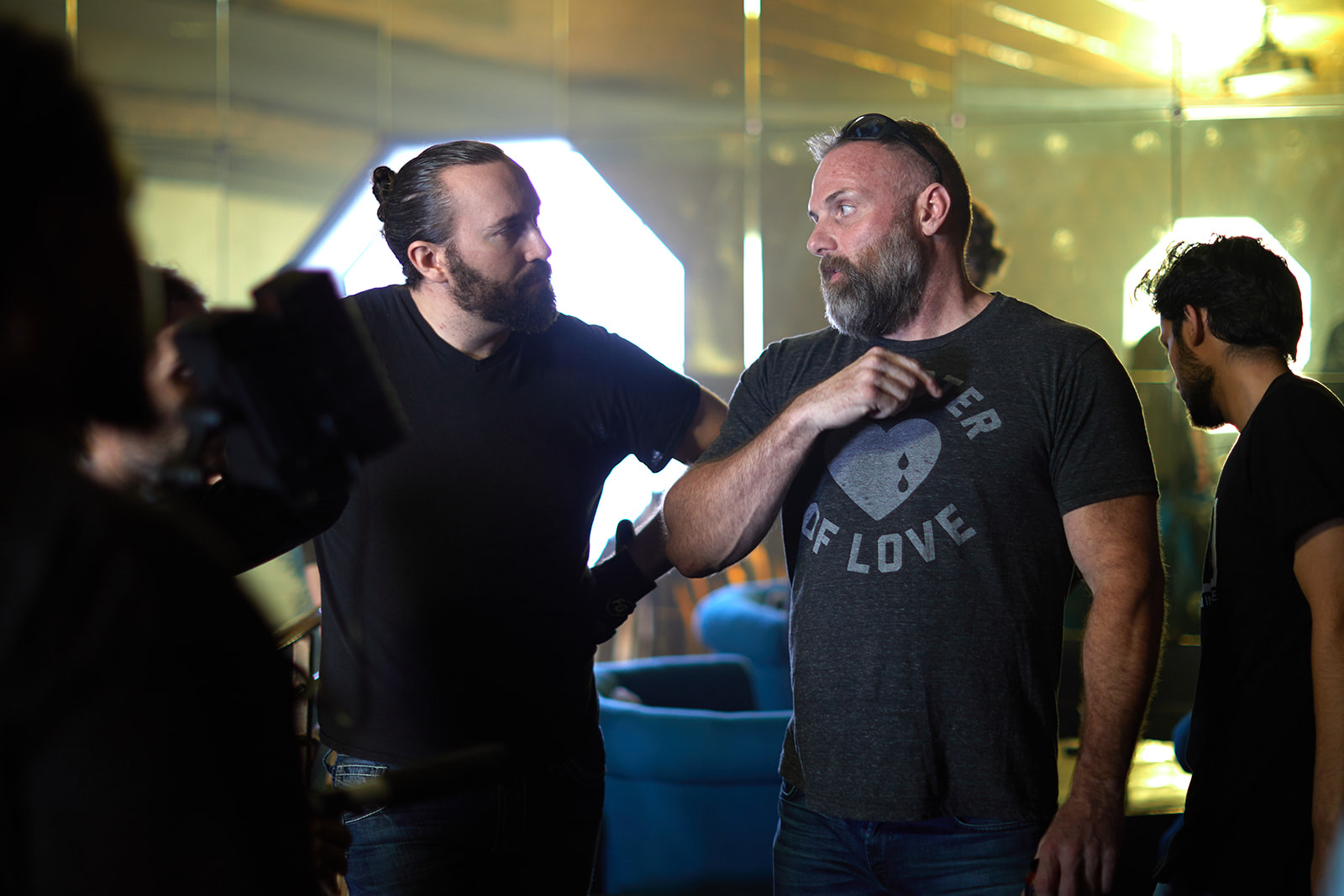 Two men with beards and long hair in conversation, one gesturing with his hand, in a dimly lit indoor setting.
