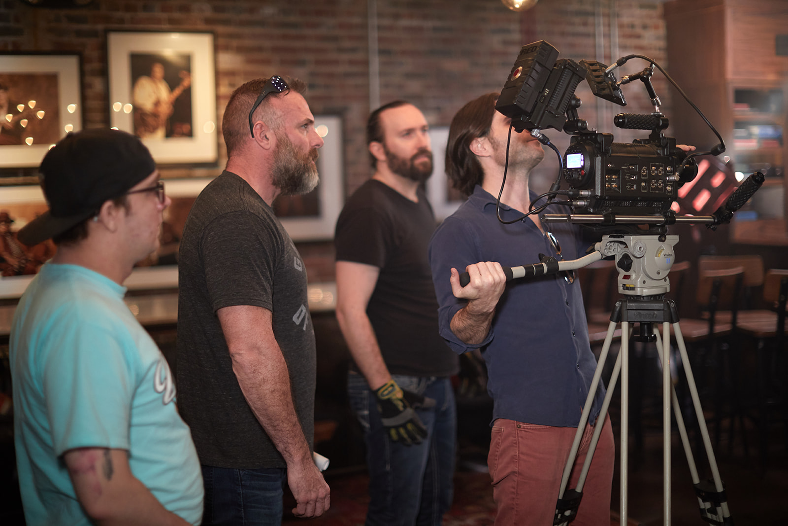 Group of four people standing in a room with a camera operator filming, brick wall background, framed pictures, and warm lighting